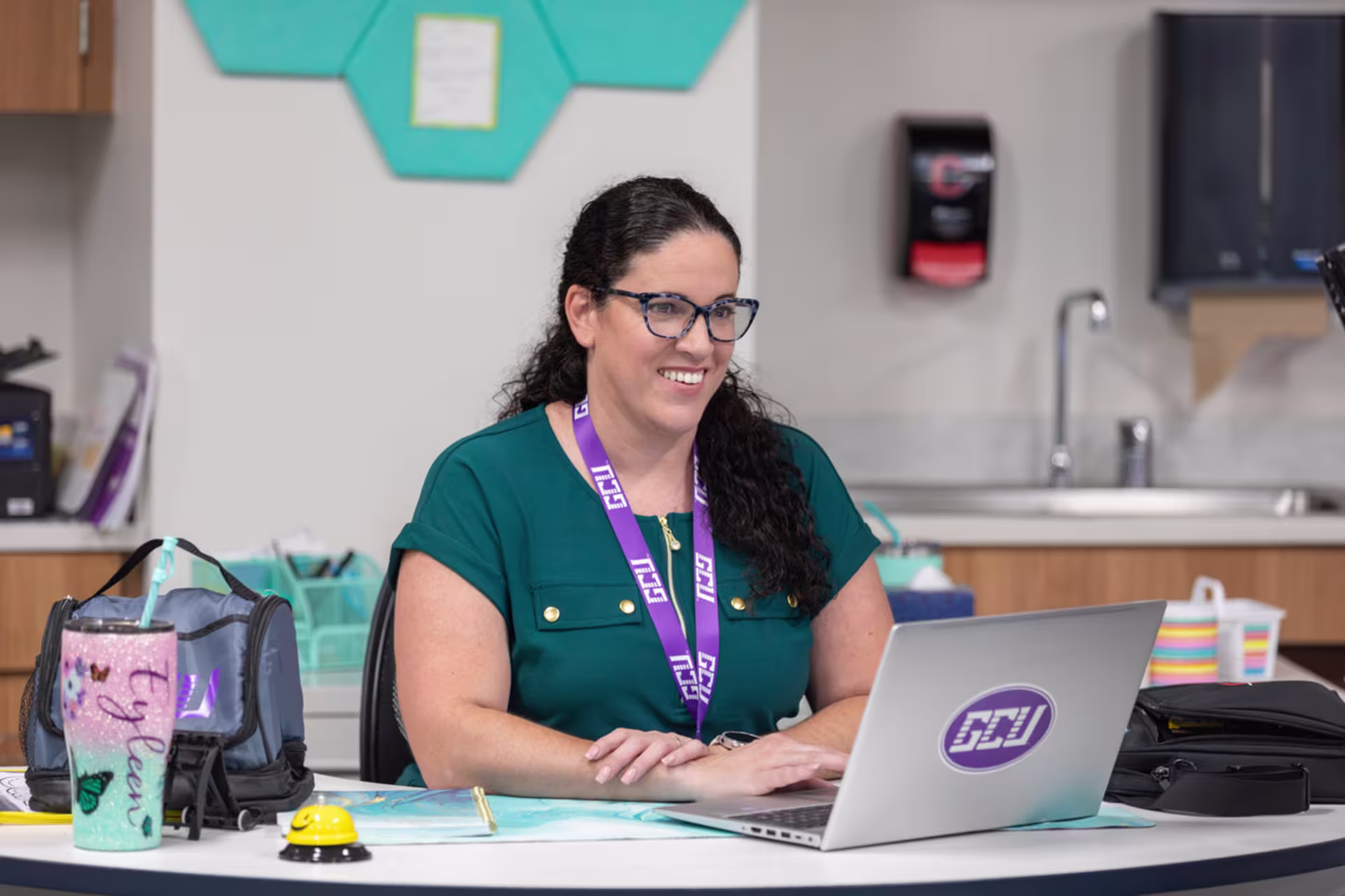 female teacher in green teal shirt wearing a GCU lanyard uses laptop for online coursework in teacher's lounge
