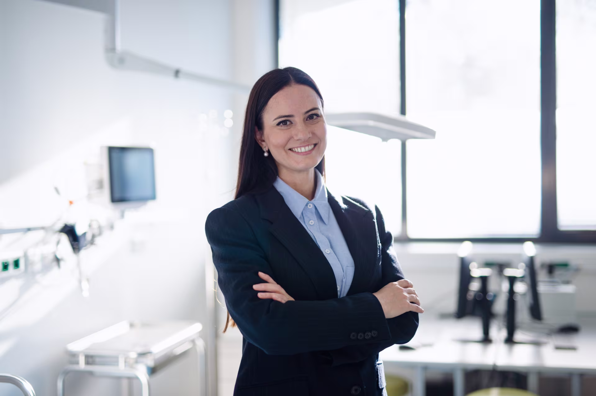 A female healthcare manager wearing a business suit with arms crossed, smiling in a hospital room.