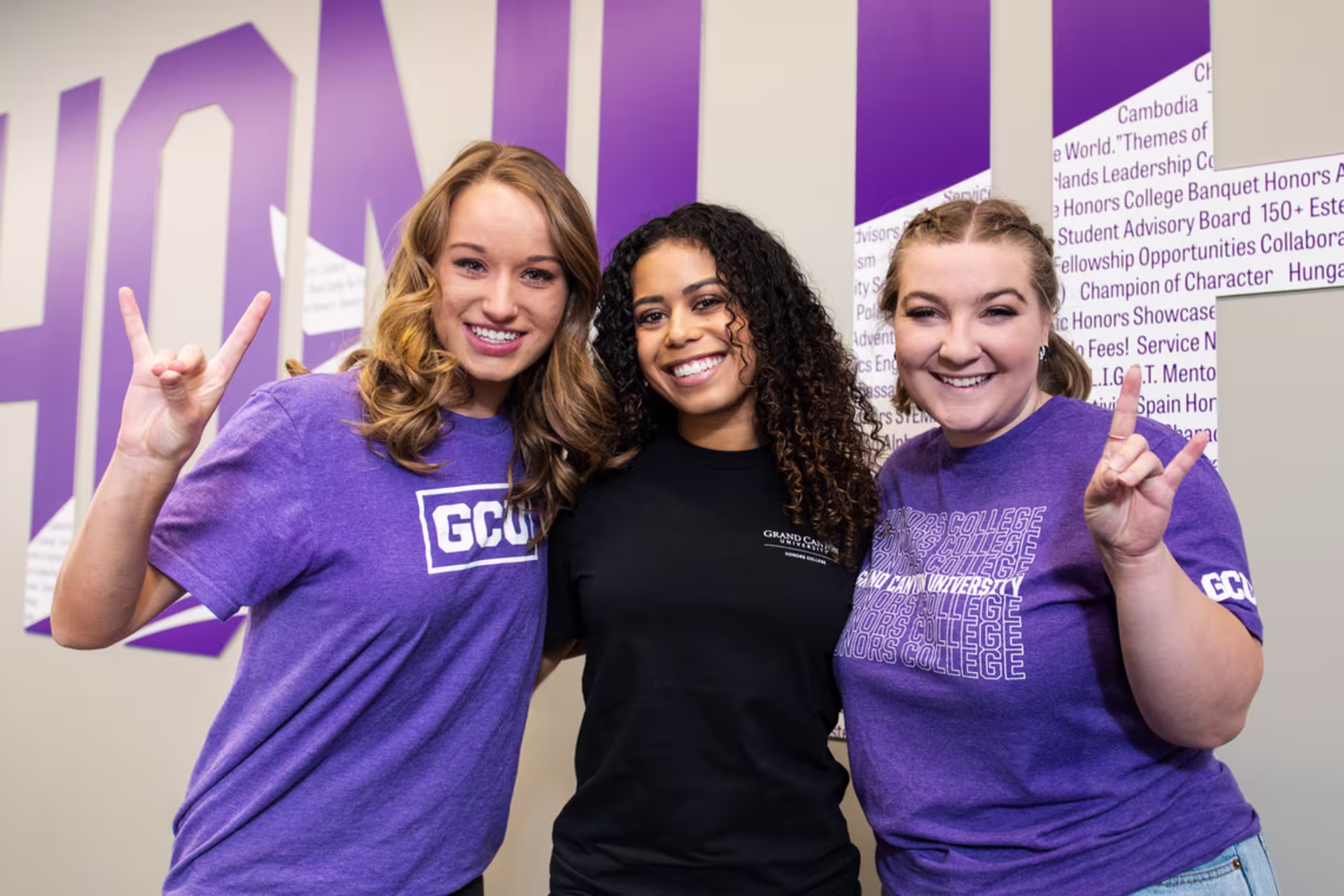 trio of female honors college students giving lopes up sign in front of honors lettering on wall