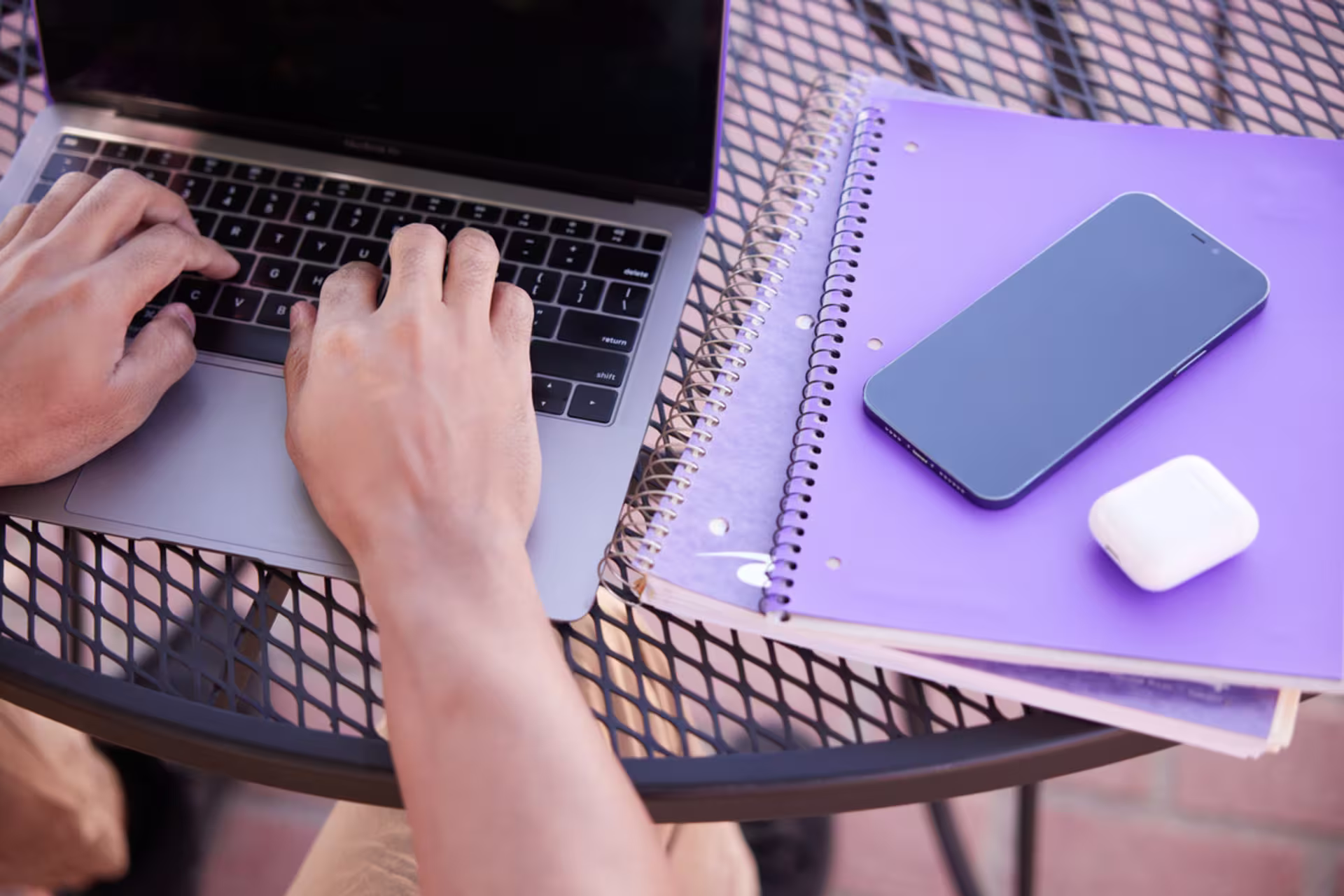 Close-up of male student using a laptop for online certificate program on outdoor table with purple notebook closed