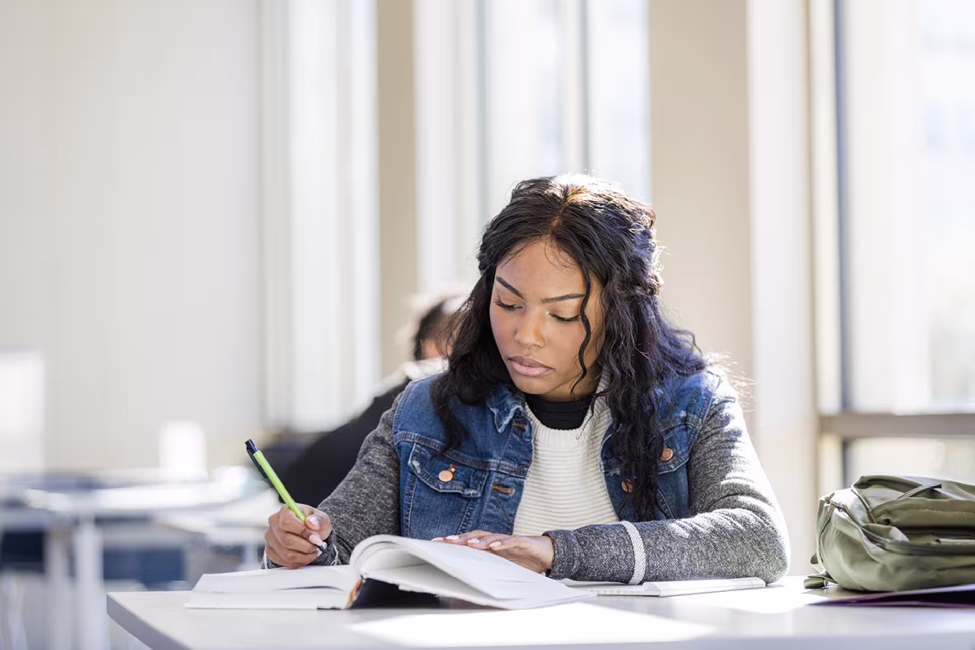 young black bachelor’s-level psychology student in jean jacket does coursework in classroom flooded by sunlight