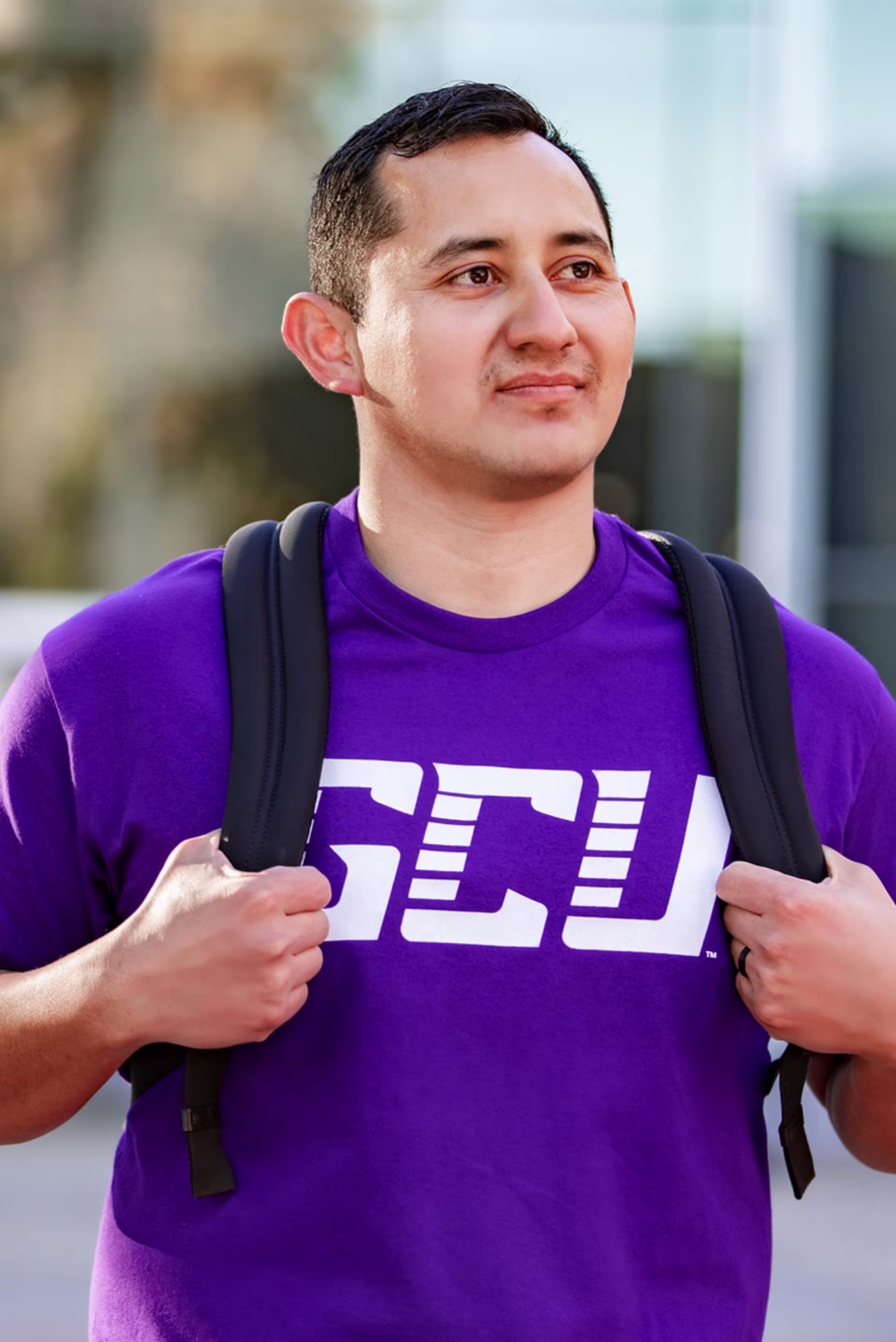 portrait image of male Hispanic student wearing a purple GCU shirt and a backpack as he walks across campus