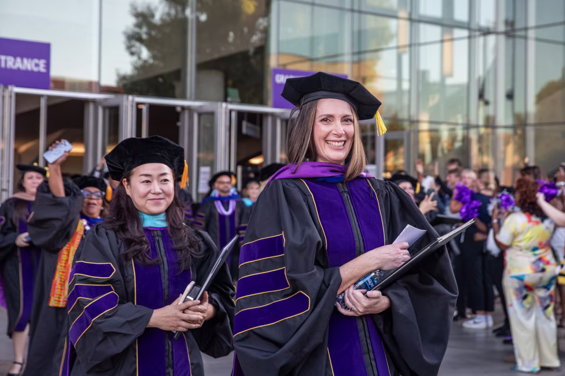 Proud brunette female doctoral graduate smiles leaving arena after GCU’s commencement followed by other doctorate graduates