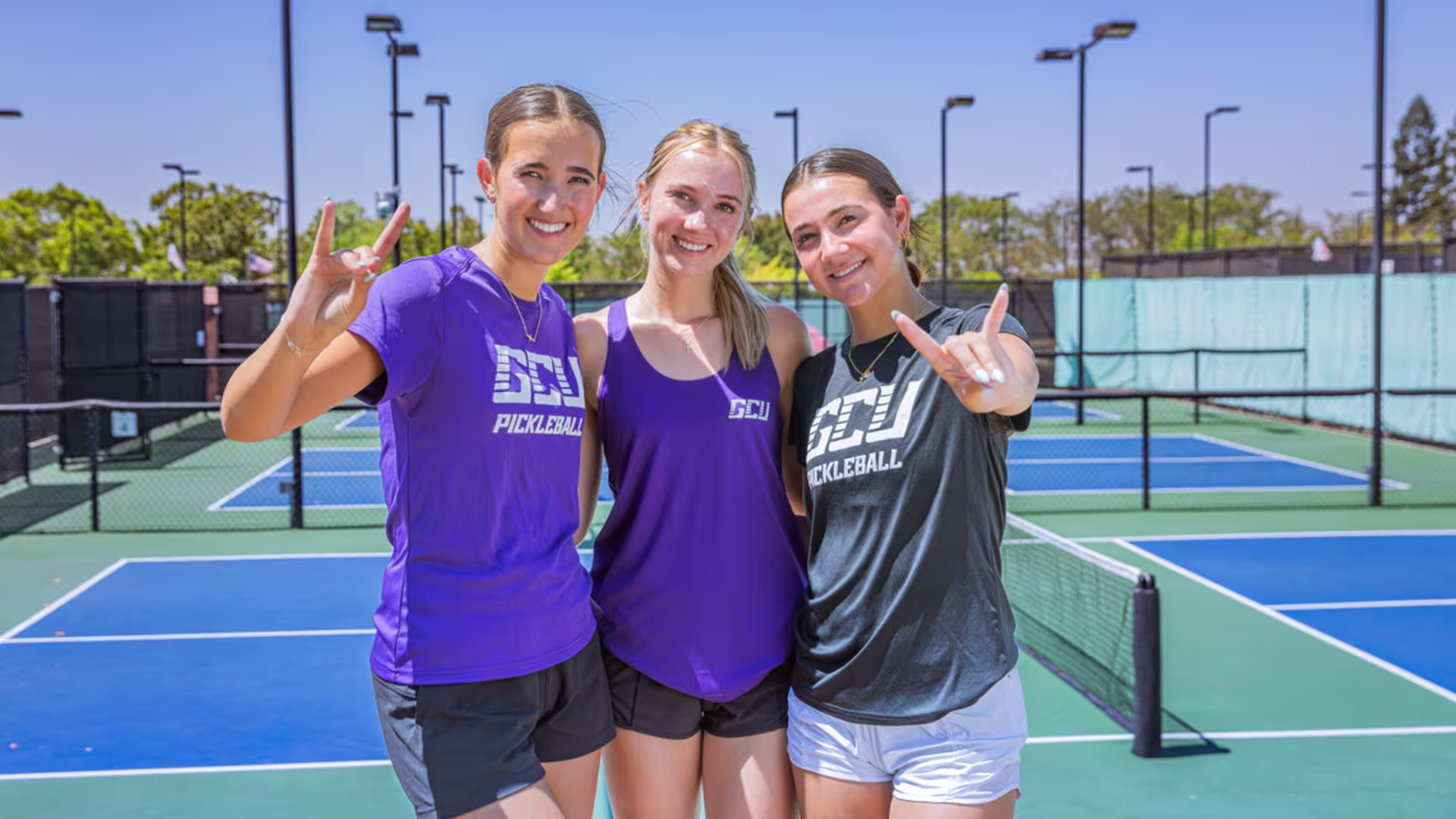 three females in GCU branded pickleball gear give lopes up sign on the campus practice court