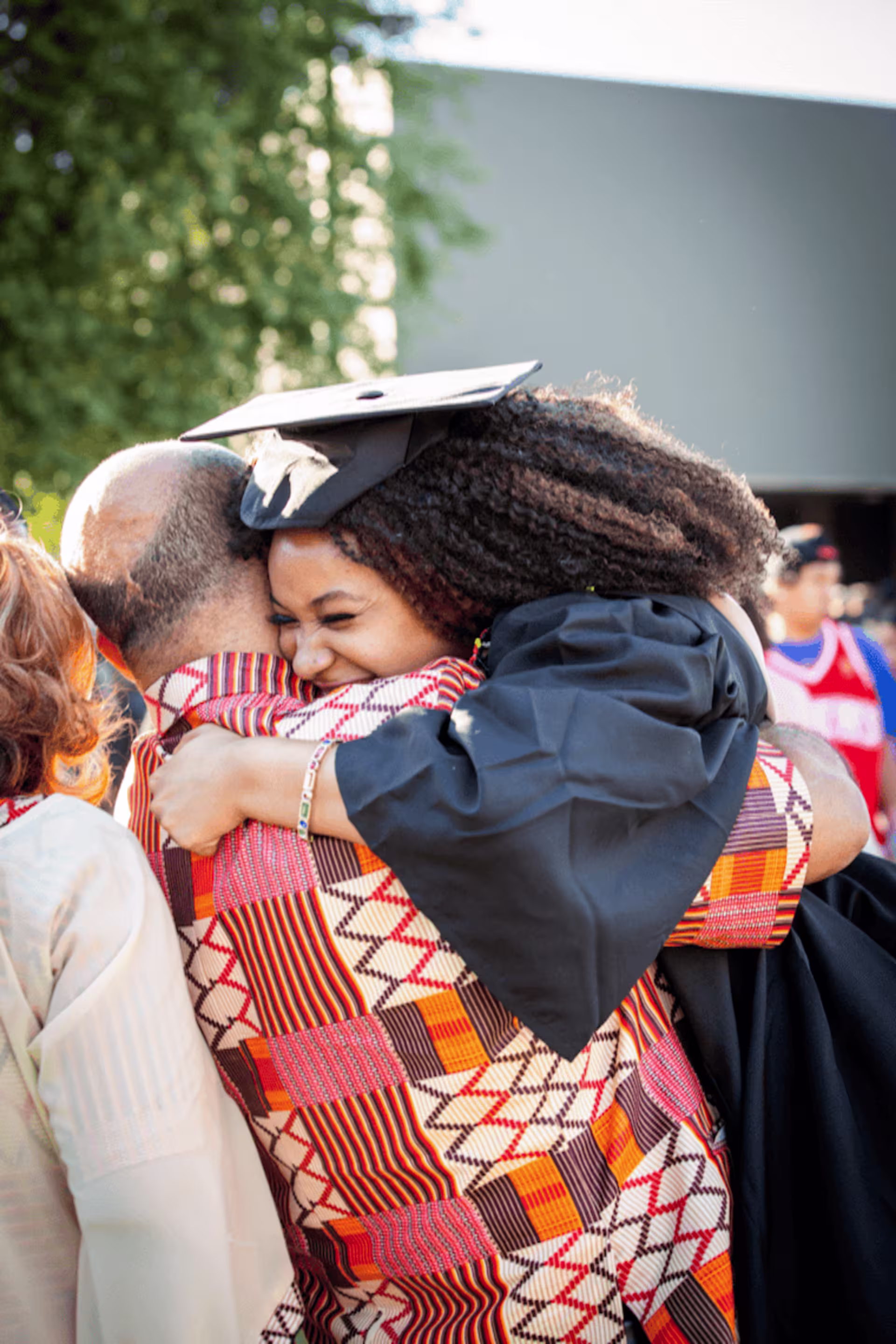 excited black female graduate hugs older man in orange patterened sweater.