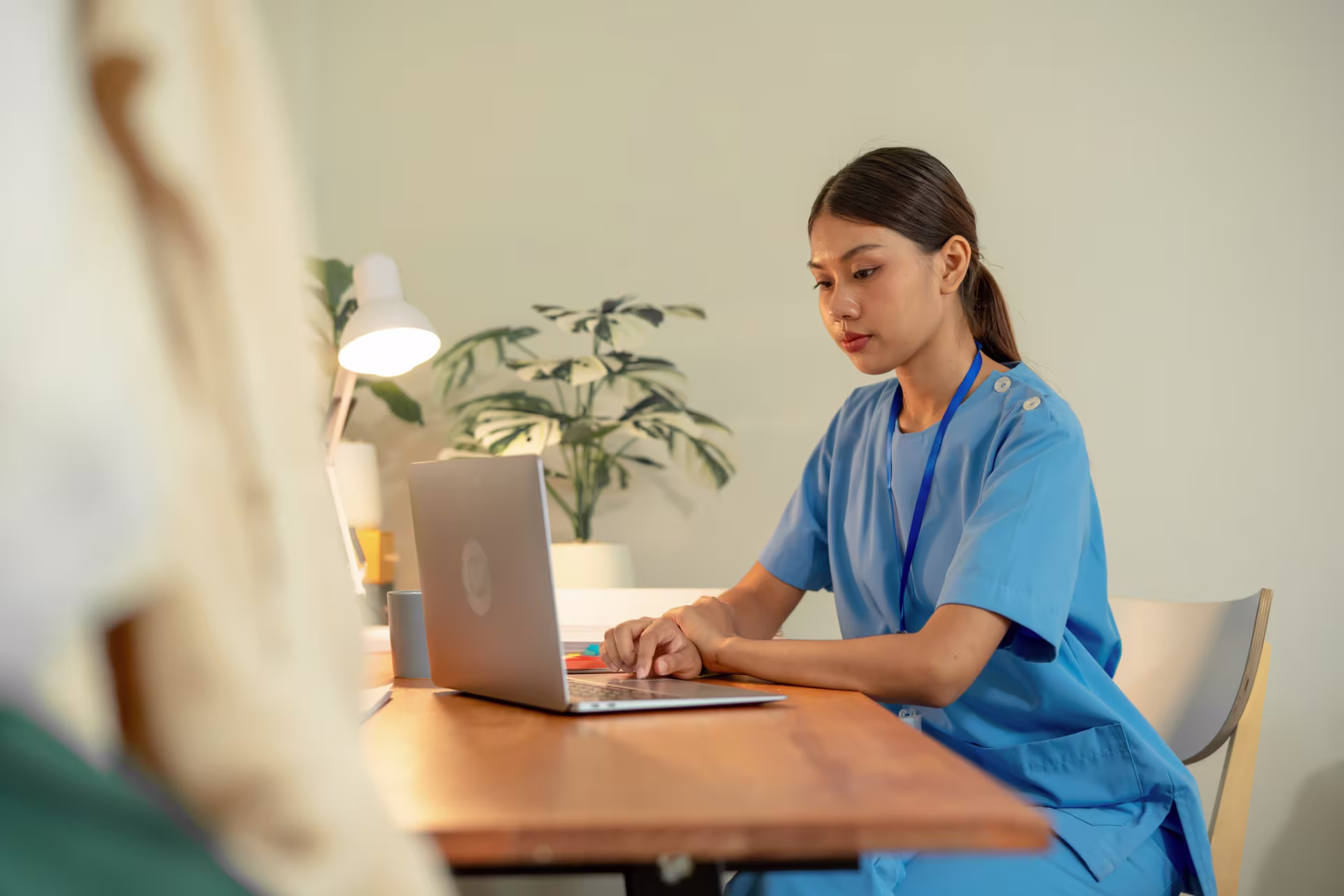 Person in blue medical scrubs typing on a laptop at a wooden desk, with a lamp and plants in the background.