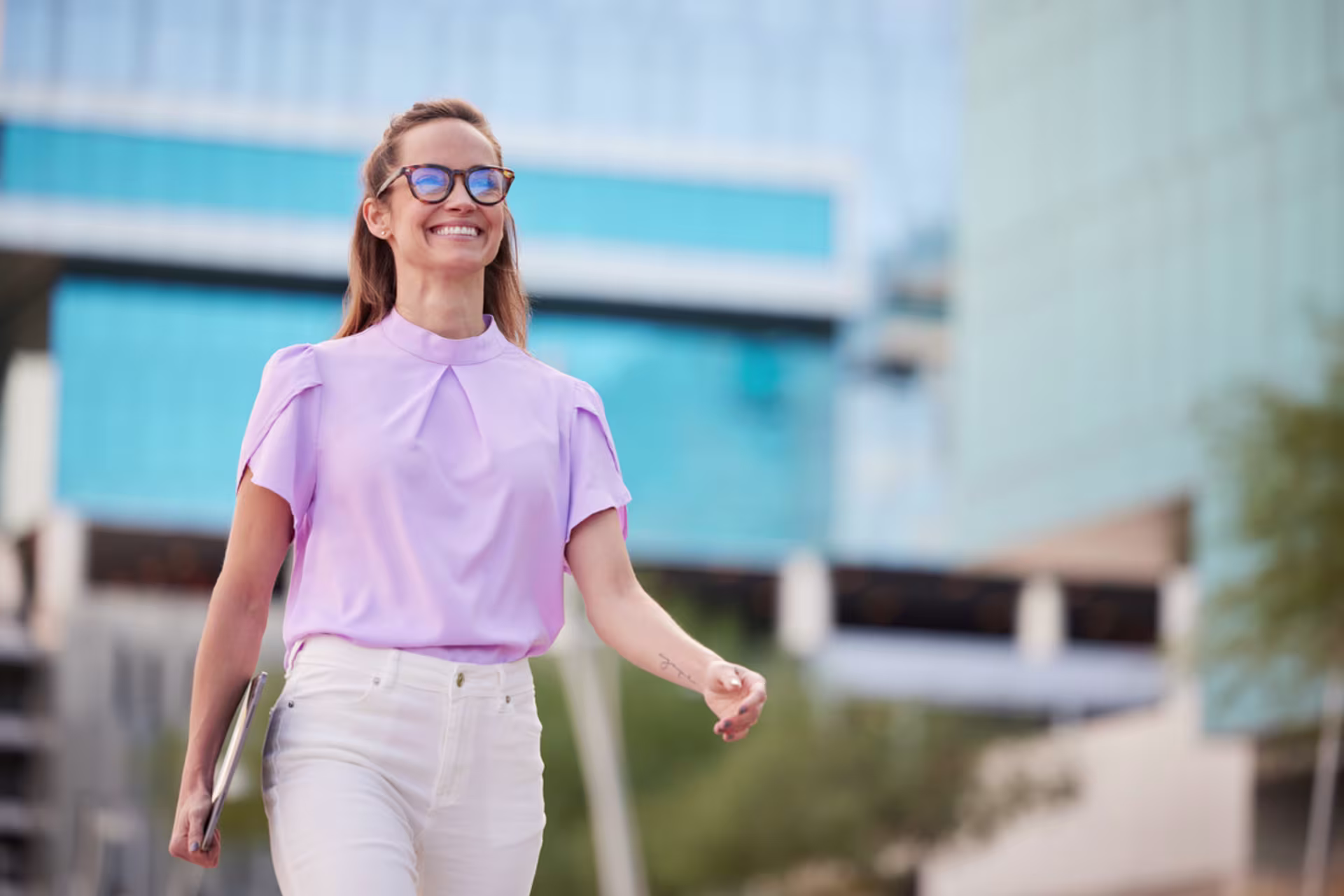 Person walking outside with a laptop, modern buildings in background.
