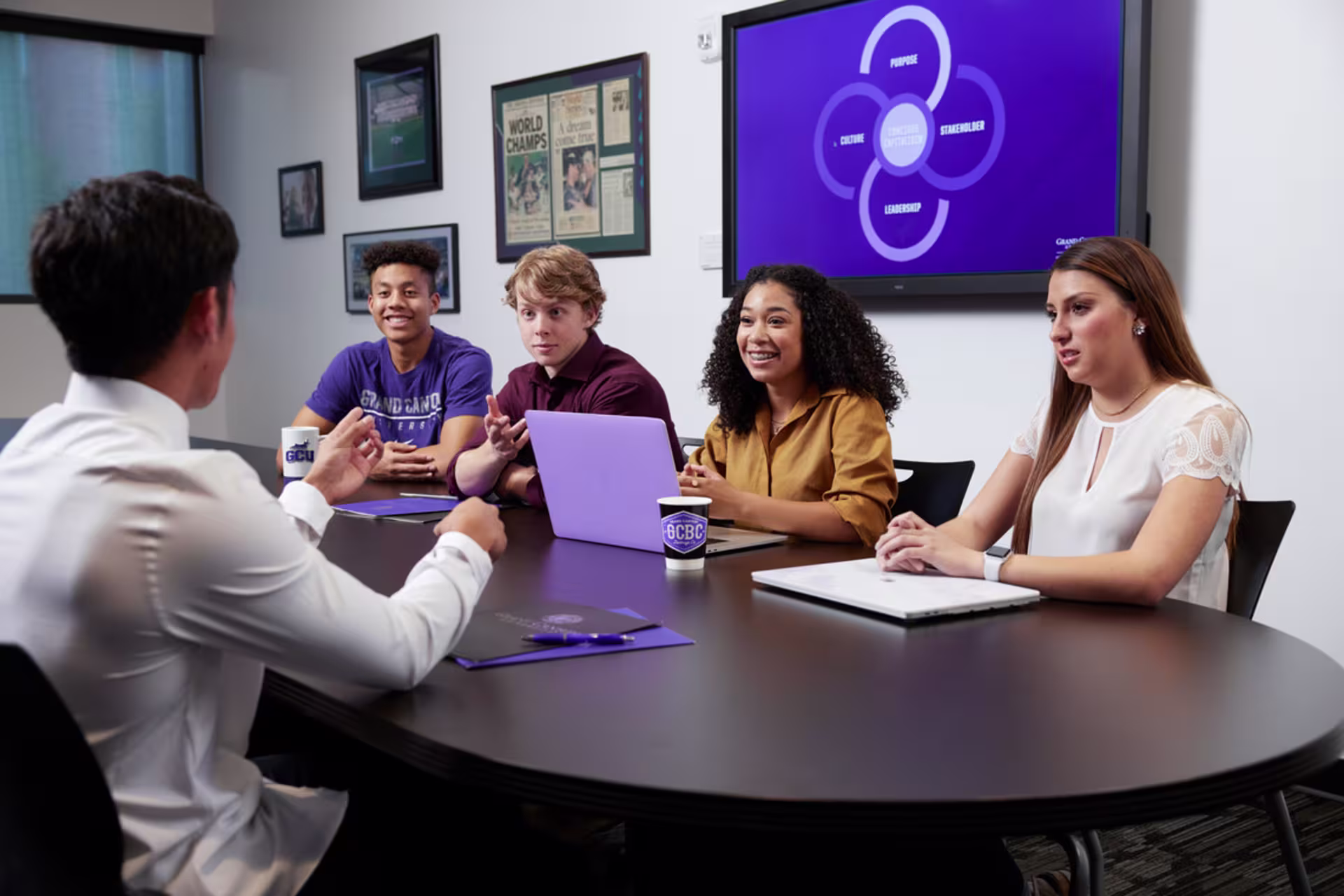 Four MBA students discuss coursework with their professor, seated around a table in a conference room.