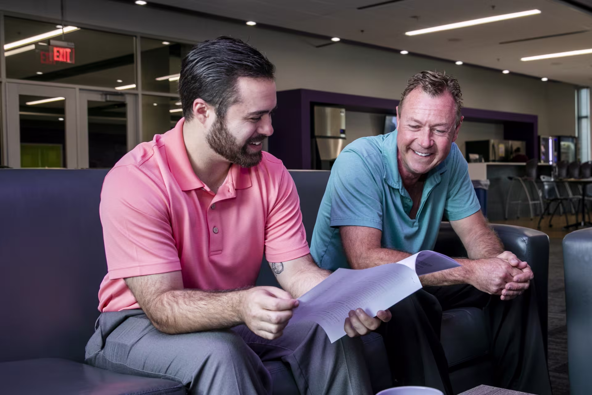 Two students sitting together in a modern lounge area, reviewing printed materials and discussing, representing collaboration in the MS in Cybersecurity Management program.