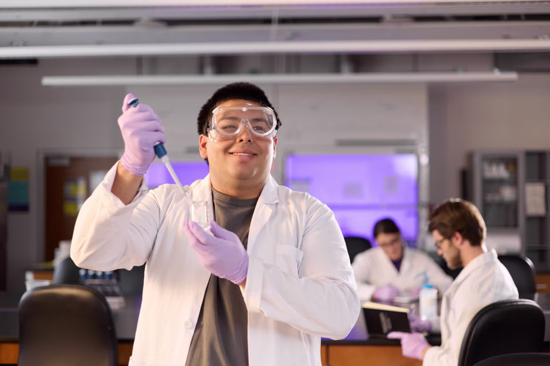 Male MLS student wearing goggles and purple gloves smiles in a lab setting while holding a pipette.