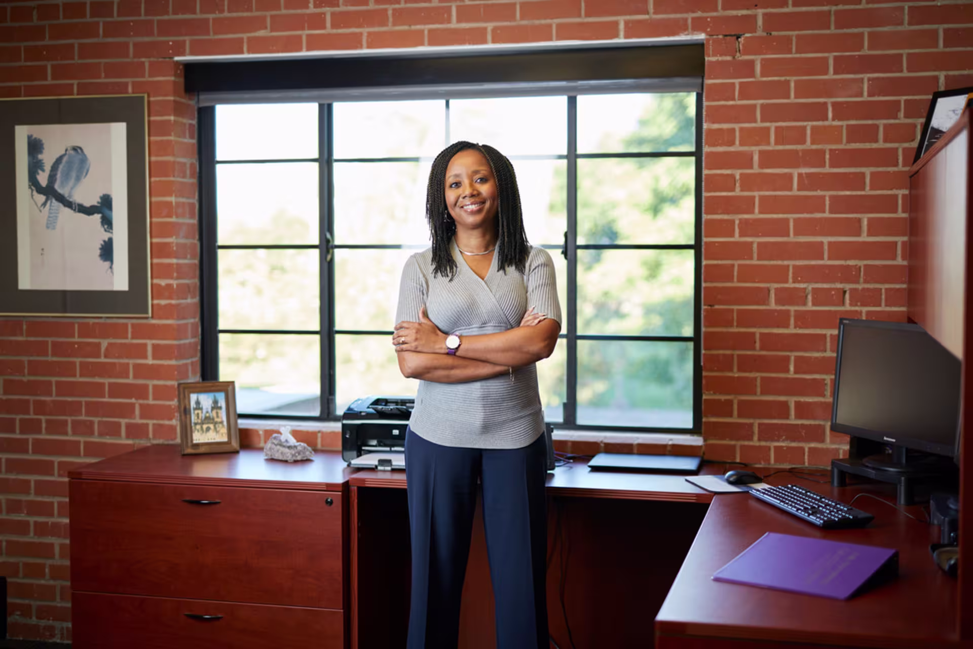 Professional woman standing confidently in an office with brick walls and large windows, arms crossed and smiling near a desk with a computer and framed photo.