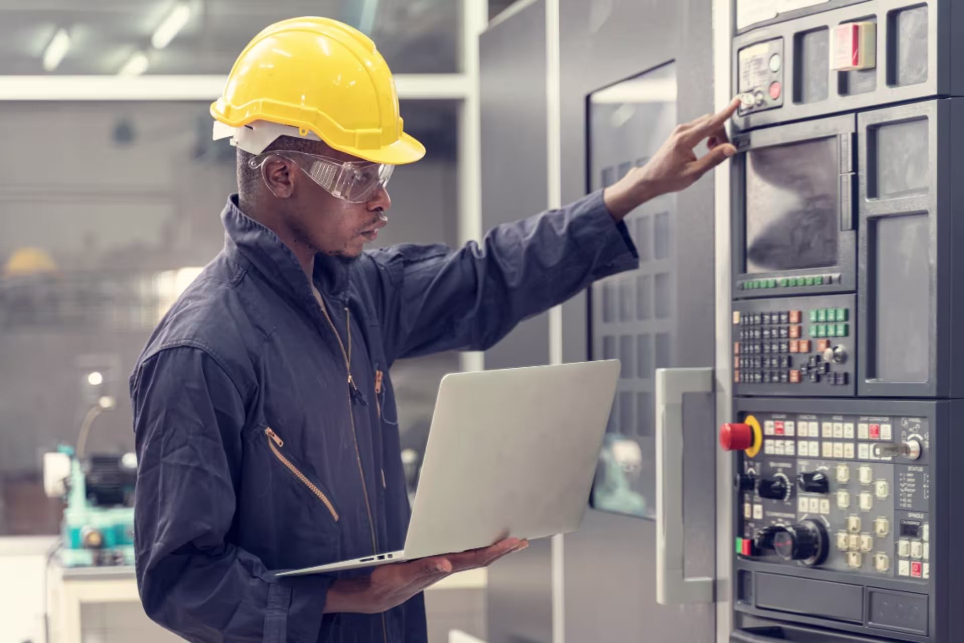 A systems engineering student wearing safety goggles, using a laptop while operating a control panel inside an engineering facility.