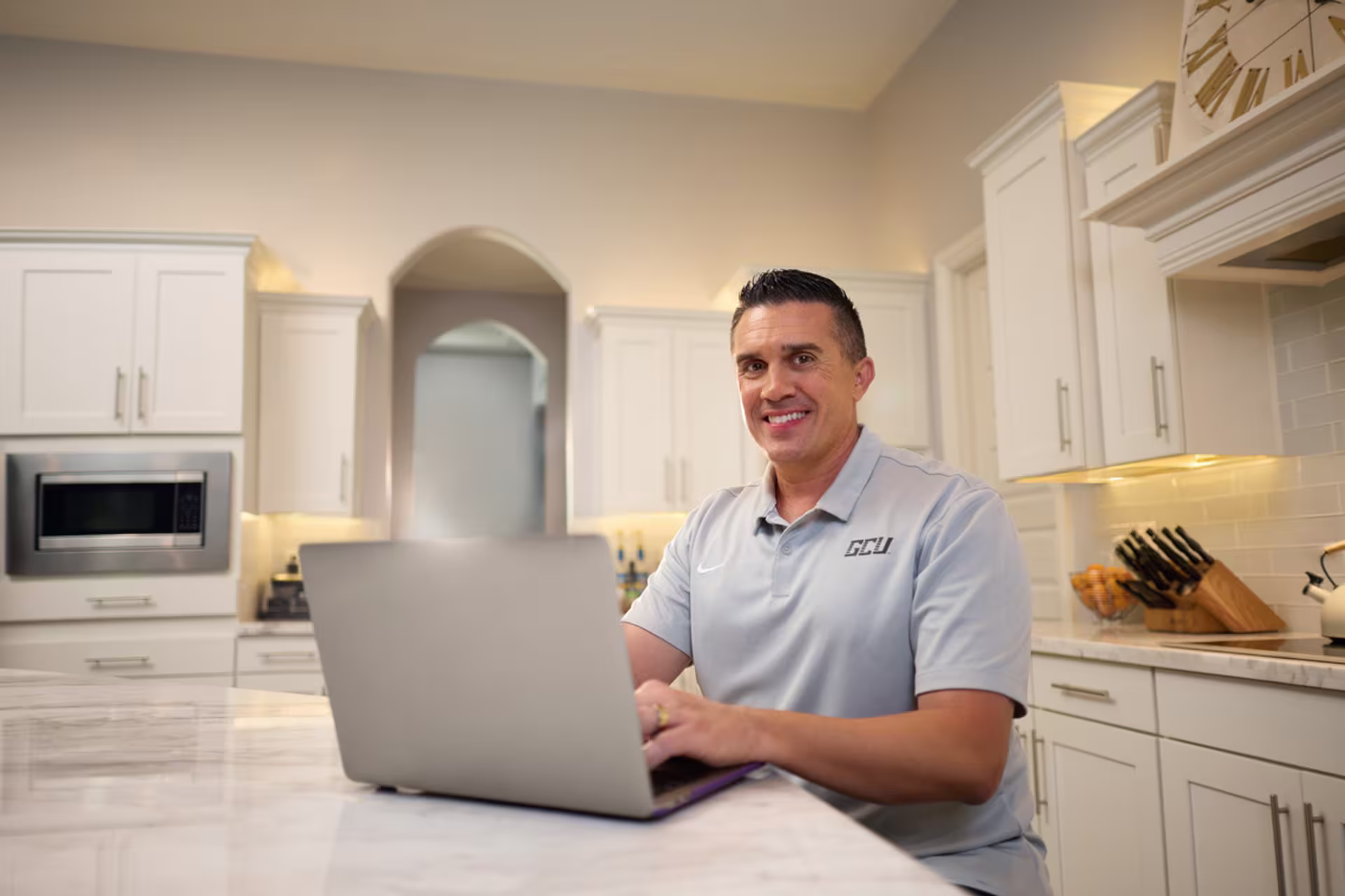 Male nonprofit MBA student sitting at a kitchen counter and smiling while typing on a laptop.