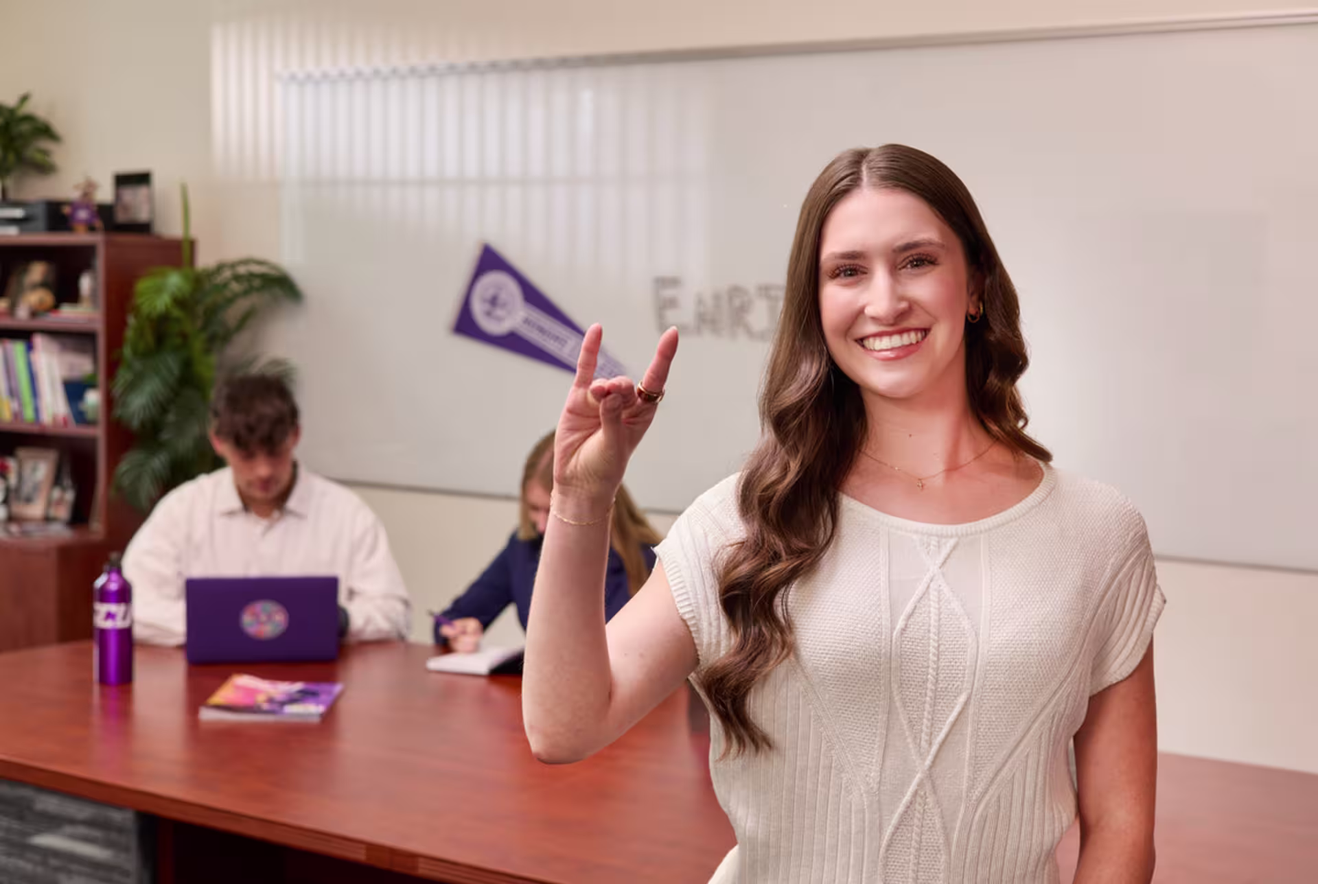 HR management student giving the lopes sign in a classroom, with two students working at a table in the background.