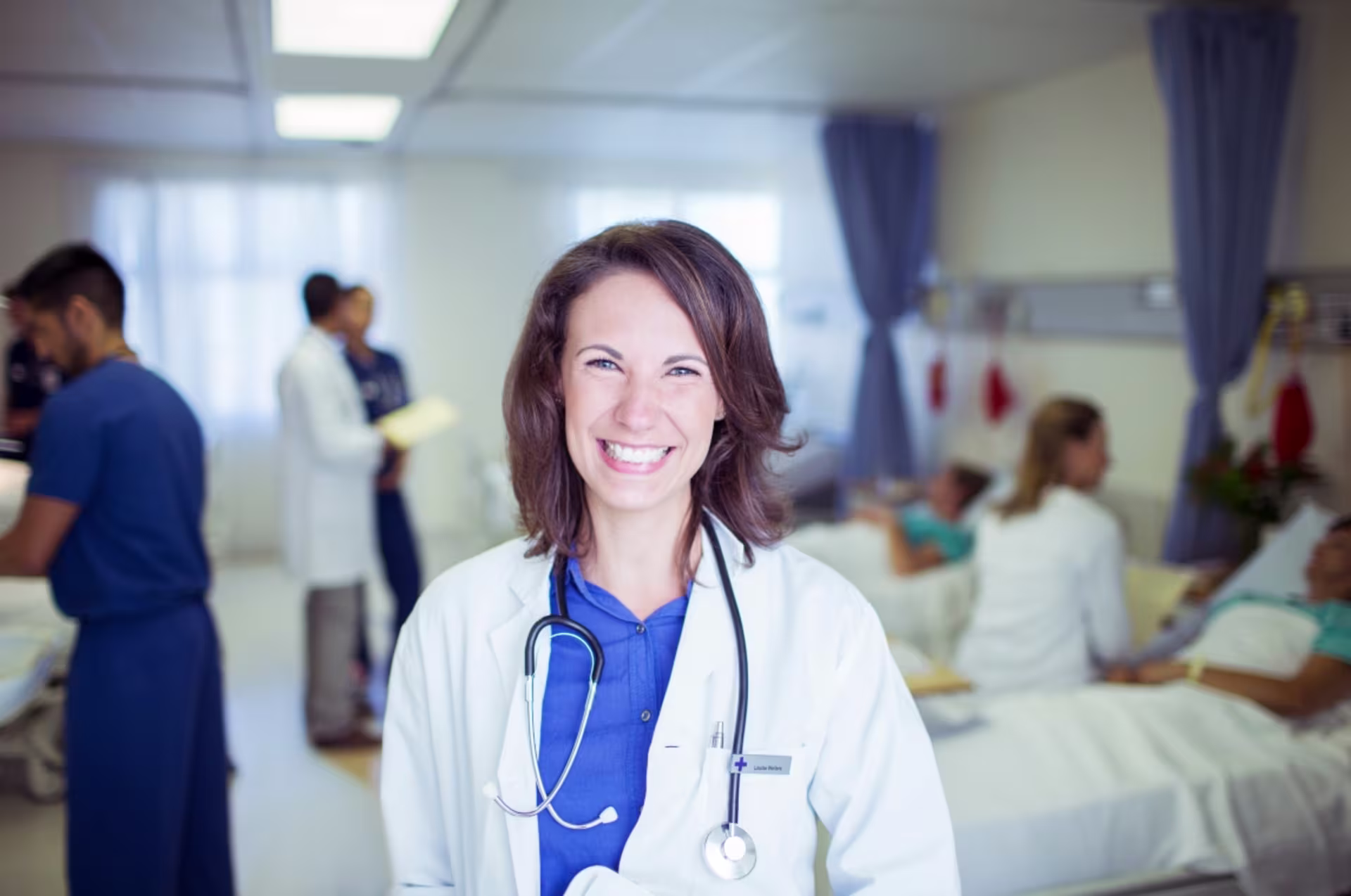 Female DNP student smiles while standing in a hospital room, with other healthcare professionals and patients in the background.