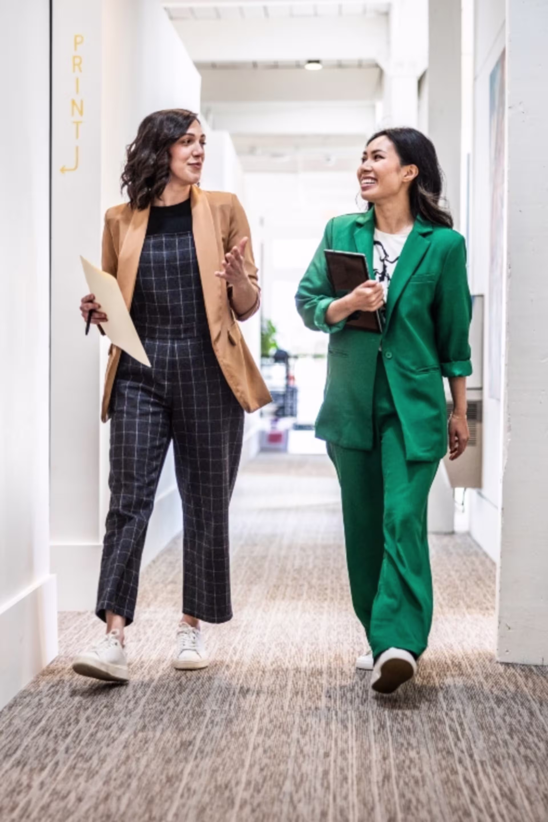Two professionals walk together down an office hallway, discussing work as they carry documents and a tablet.