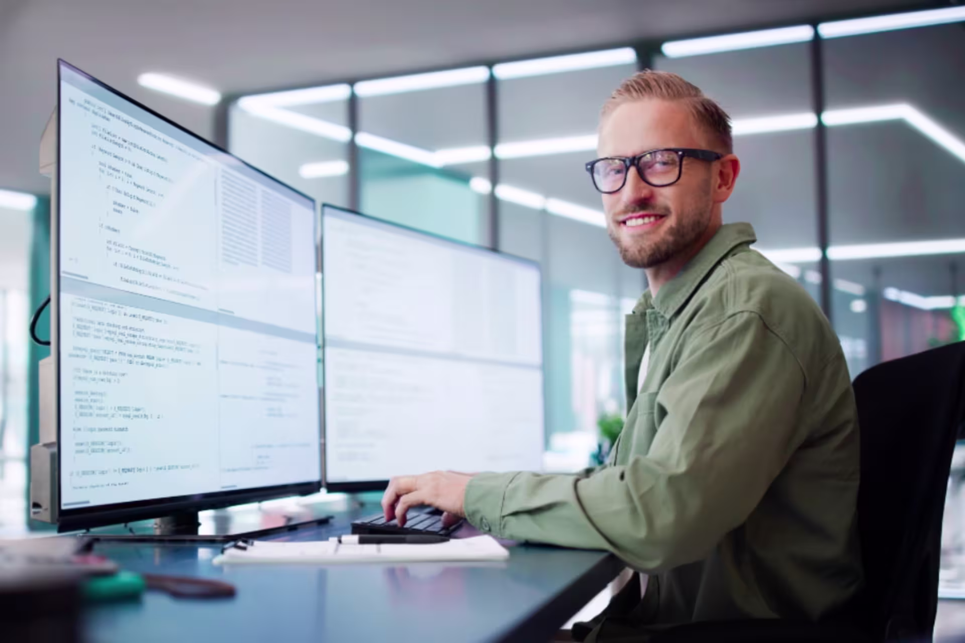 A professional developer works at a desk with dual monitors displaying code in a modern office environment.