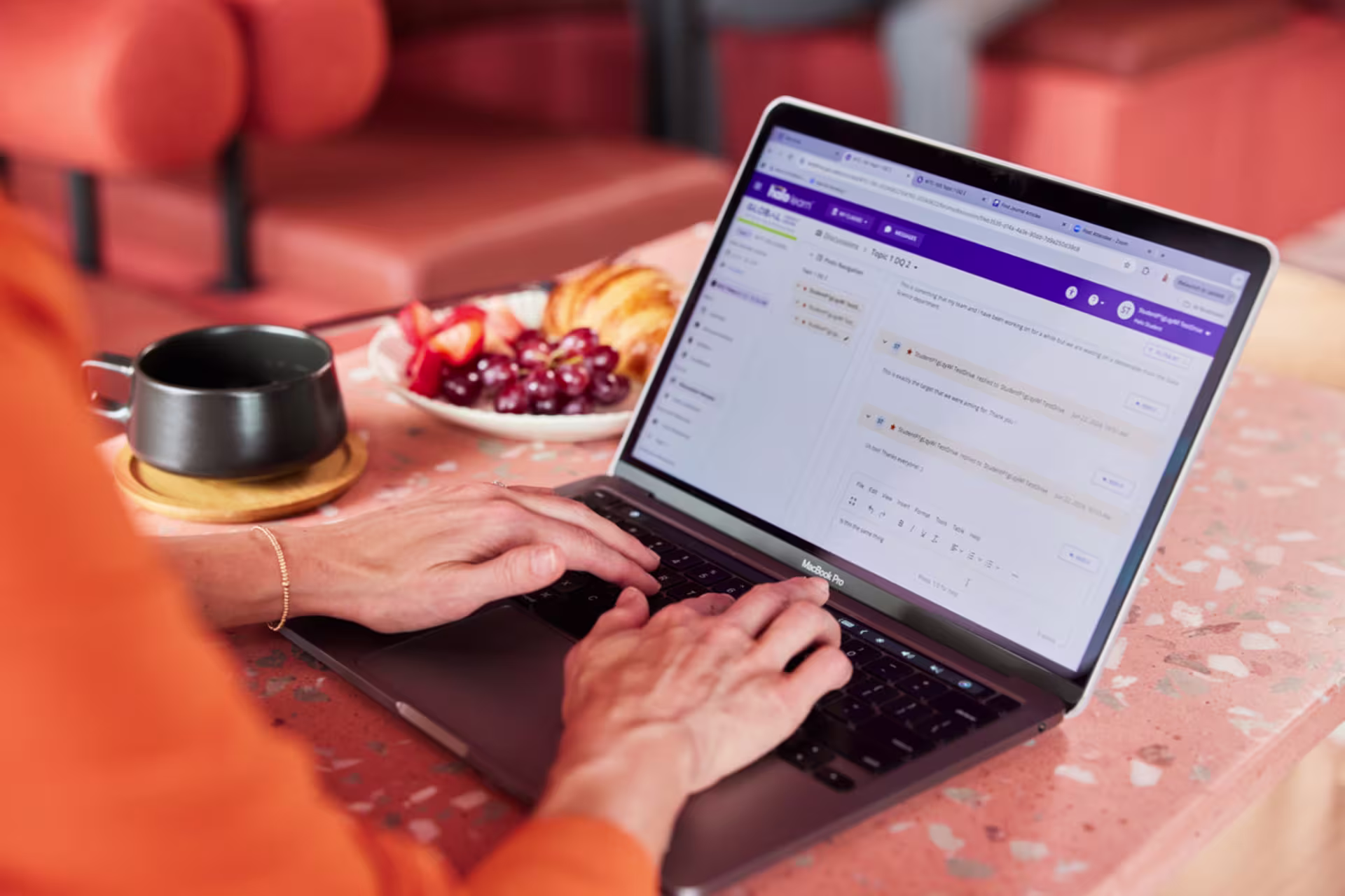 A person works on a laptop at a café-style table with online coursework displayed on the screen, alongside a cup of coffee and a plate of fruit.
