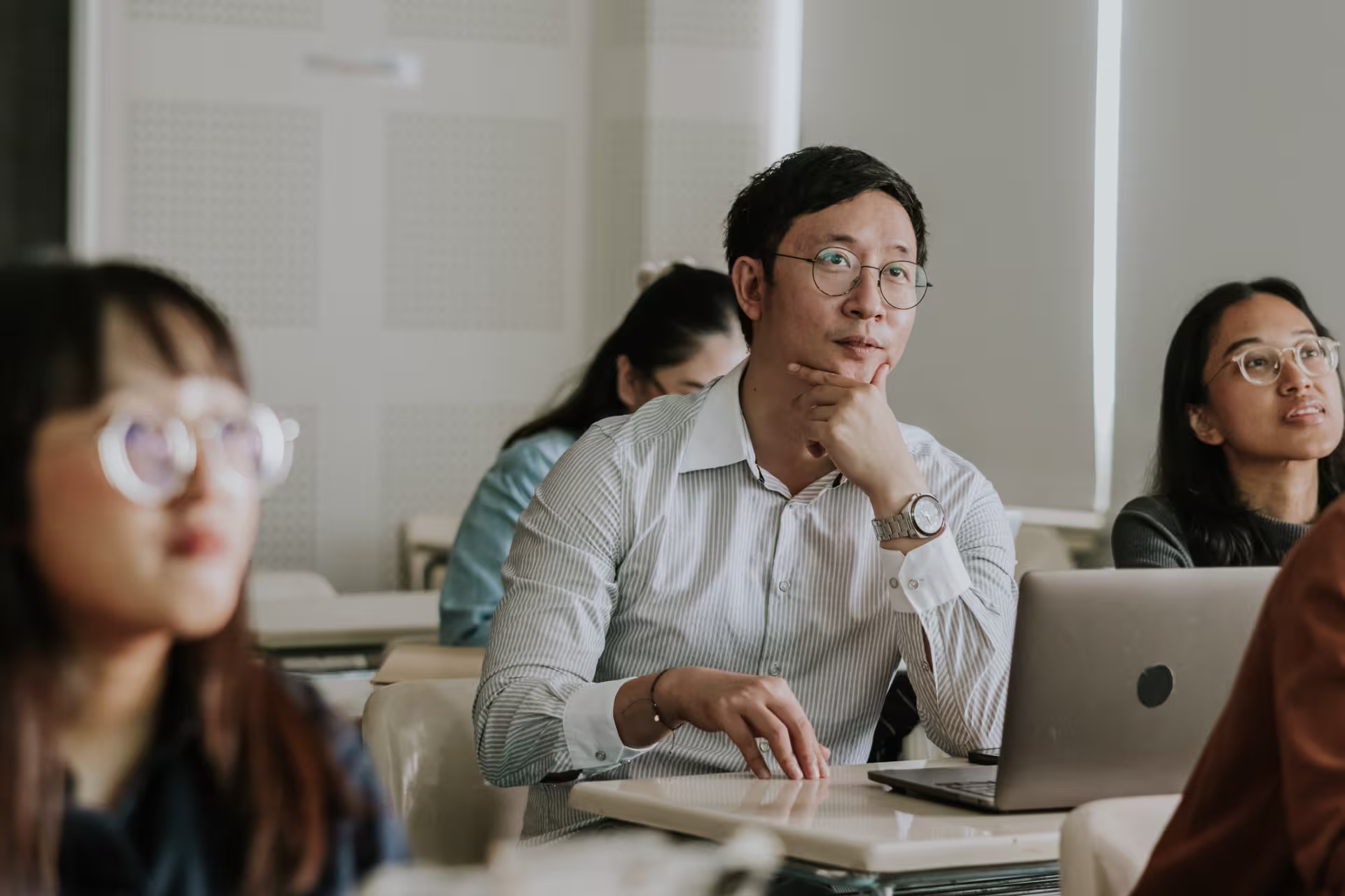 A group of students sits in a classroom, with one person using a laptop while listening attentively.
