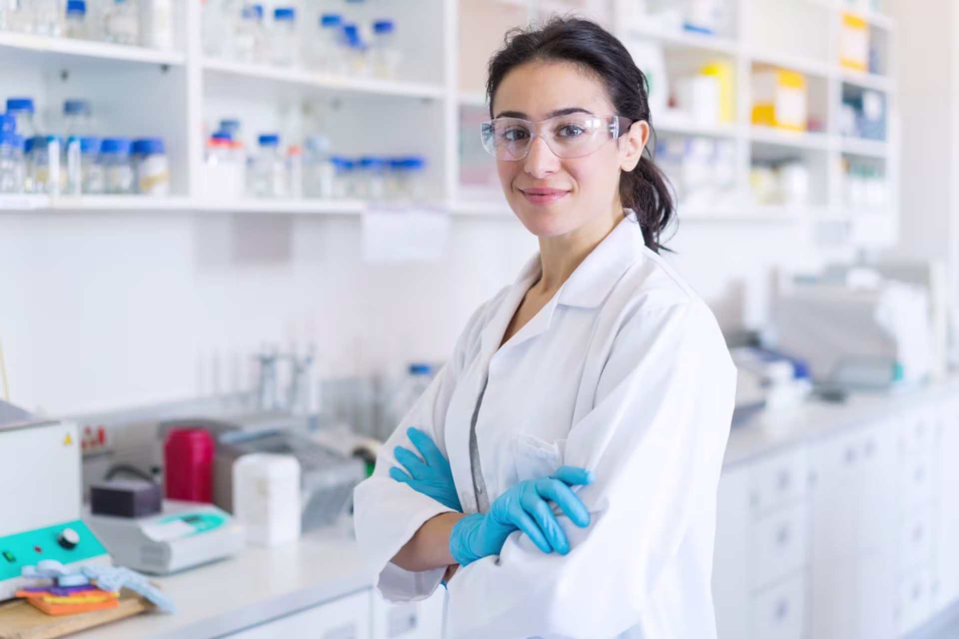 Female biomedical engineering student wearing a lab coat, safety goggles, and gloves standing in a lab with equipment in the background.