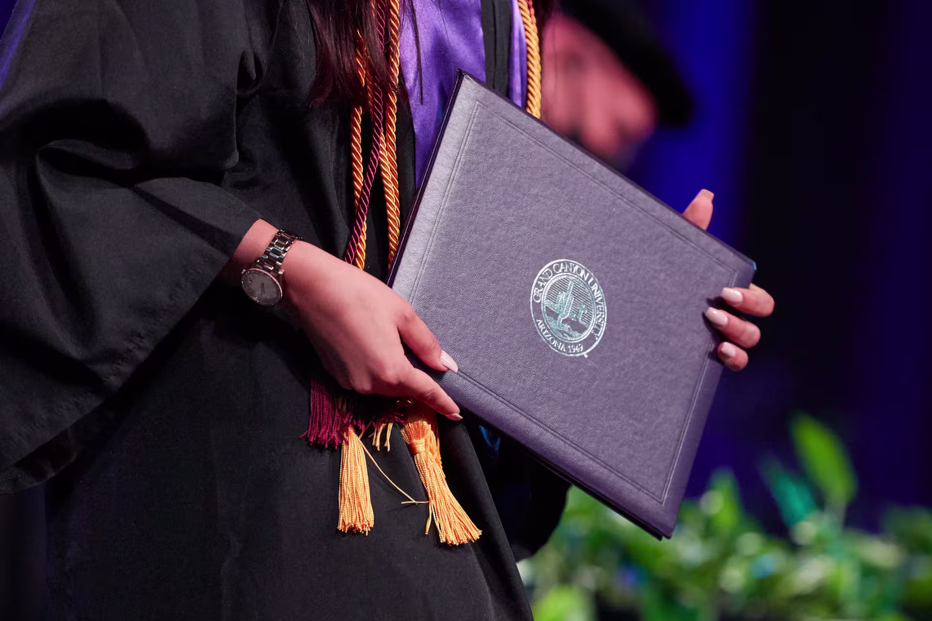 Graduate wearing a cap and gown holds a Grand Canyon University diploma cover during a commencement ceremony.
