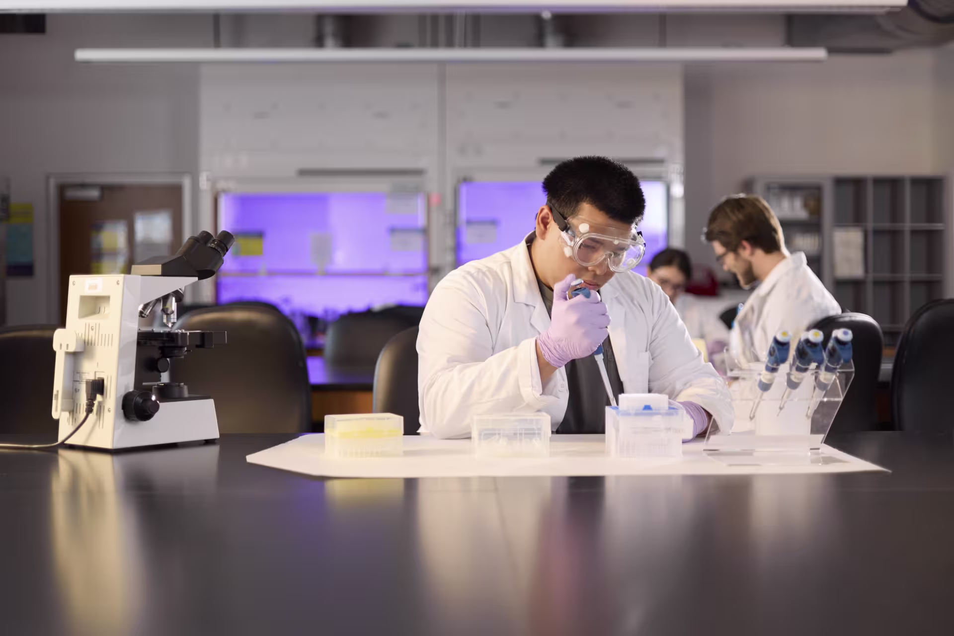Male MS Biology student in a lab wearing a lab coat and goggles.