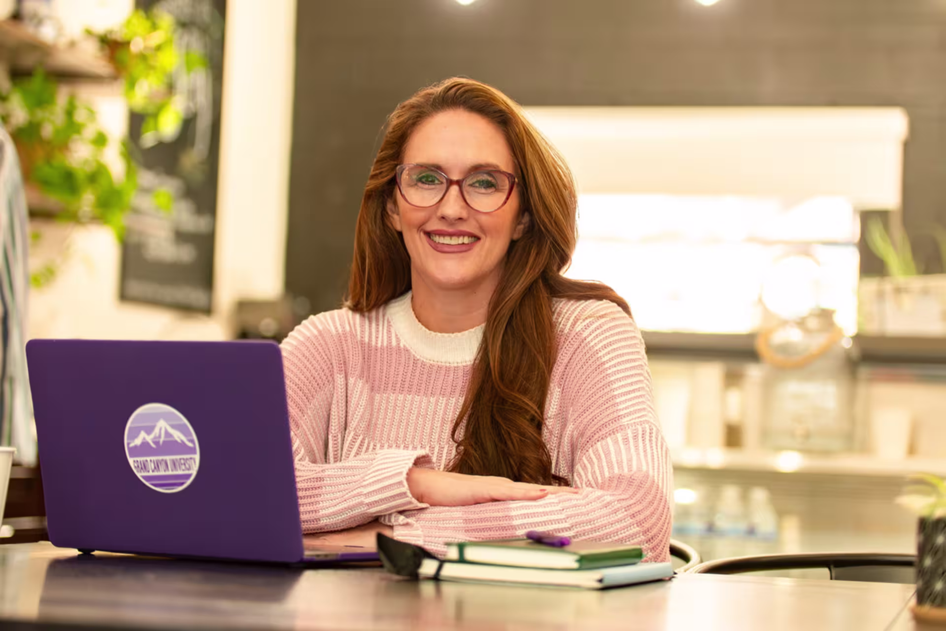 A woman sits at a table with a laptop and notebooks, smiling while working in a bright, casual study environment.