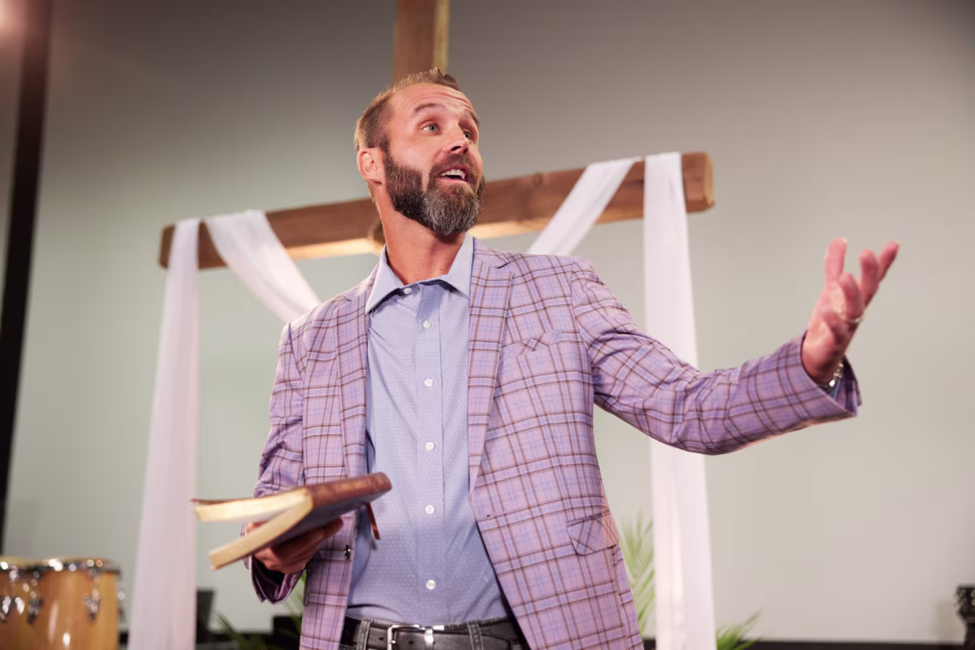 A male New Testament studies professional holding an open Bible while speaking in front of a wooden cross.