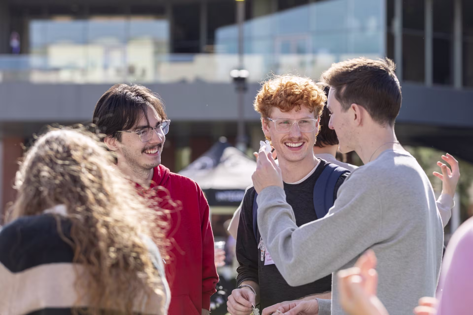 A group of students gathers outdoors on a sunny day, talking and interacting near a campus building.