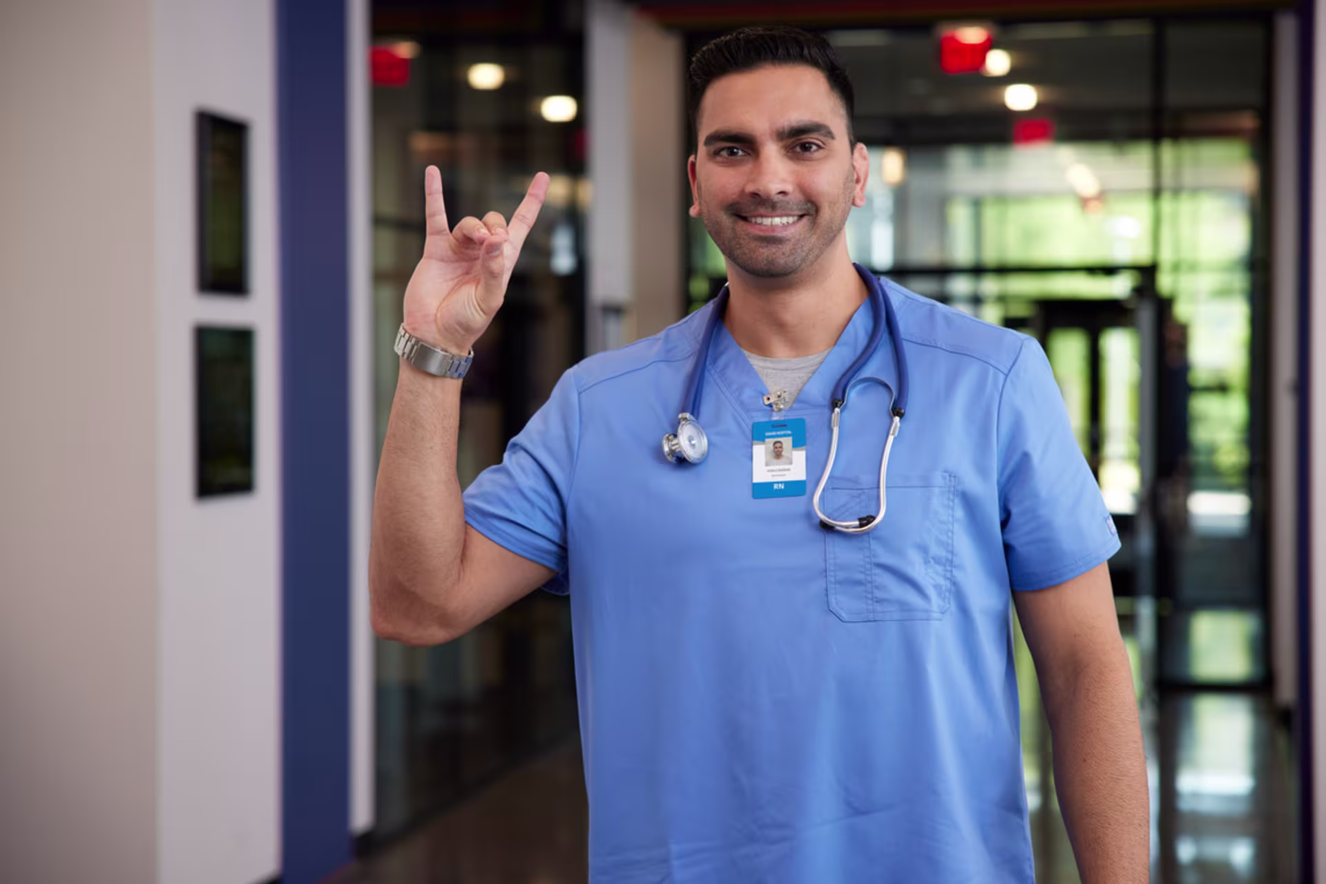 Healthcare worker in scrubs standing in a medical facility, raising a lopes up gesture while wearing a stethoscope.