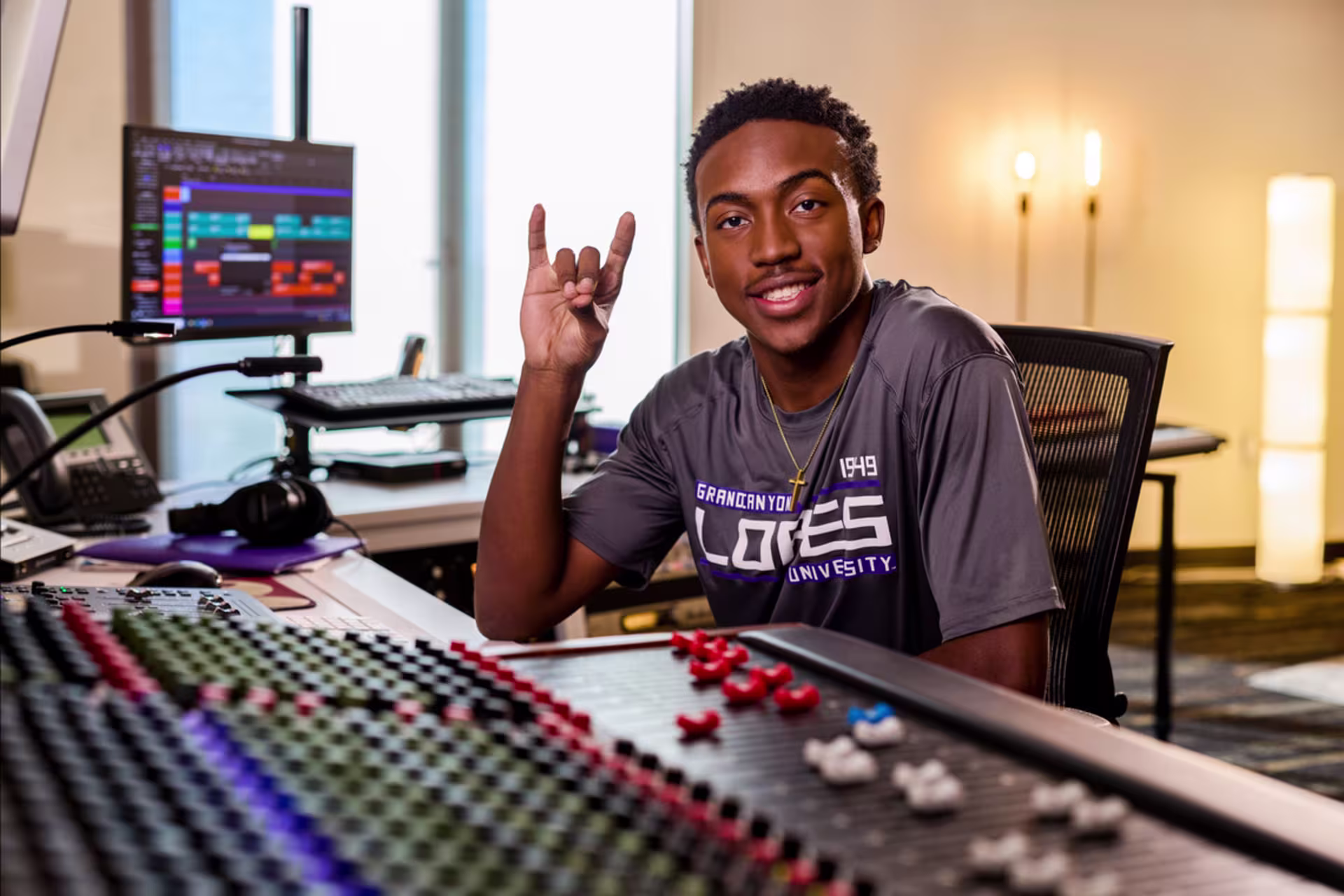 Student working at an audio mixing console in a media production studio, raising lopes up while monitoring sound levels.