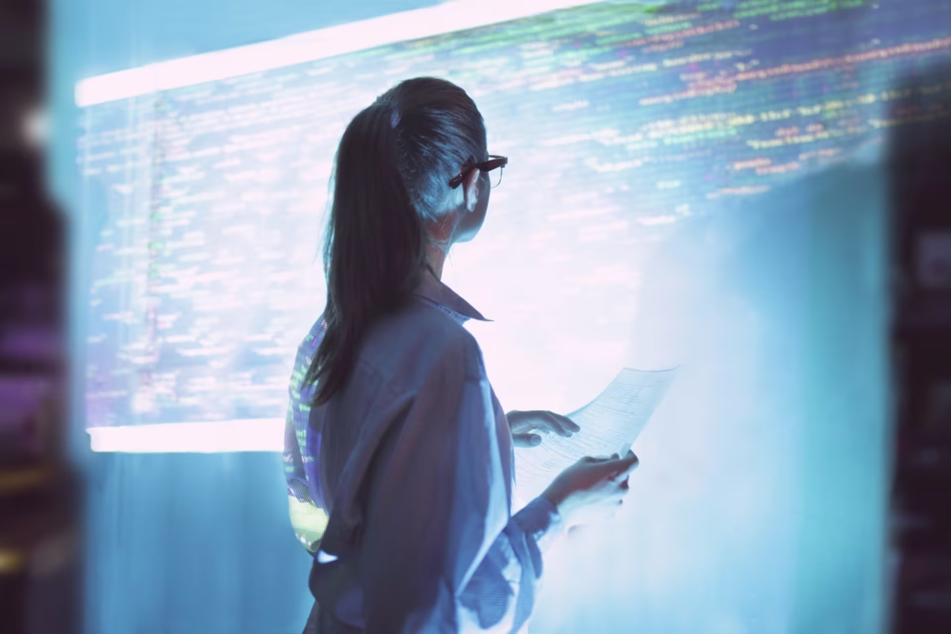 Woman reviewing digital code on a large screen while holding documents in a modern technology workspace.