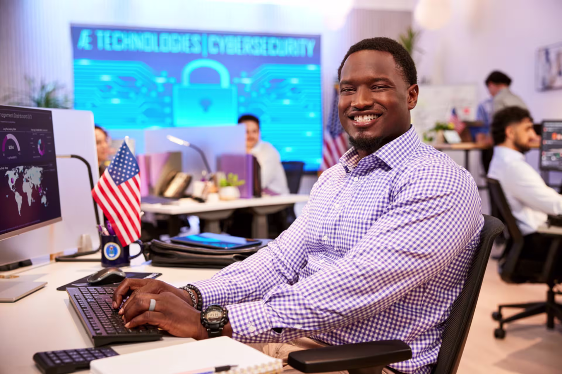 Smiling information security analyst working at a computer in a modern office with digital security graphics displayed on a screen behind him.