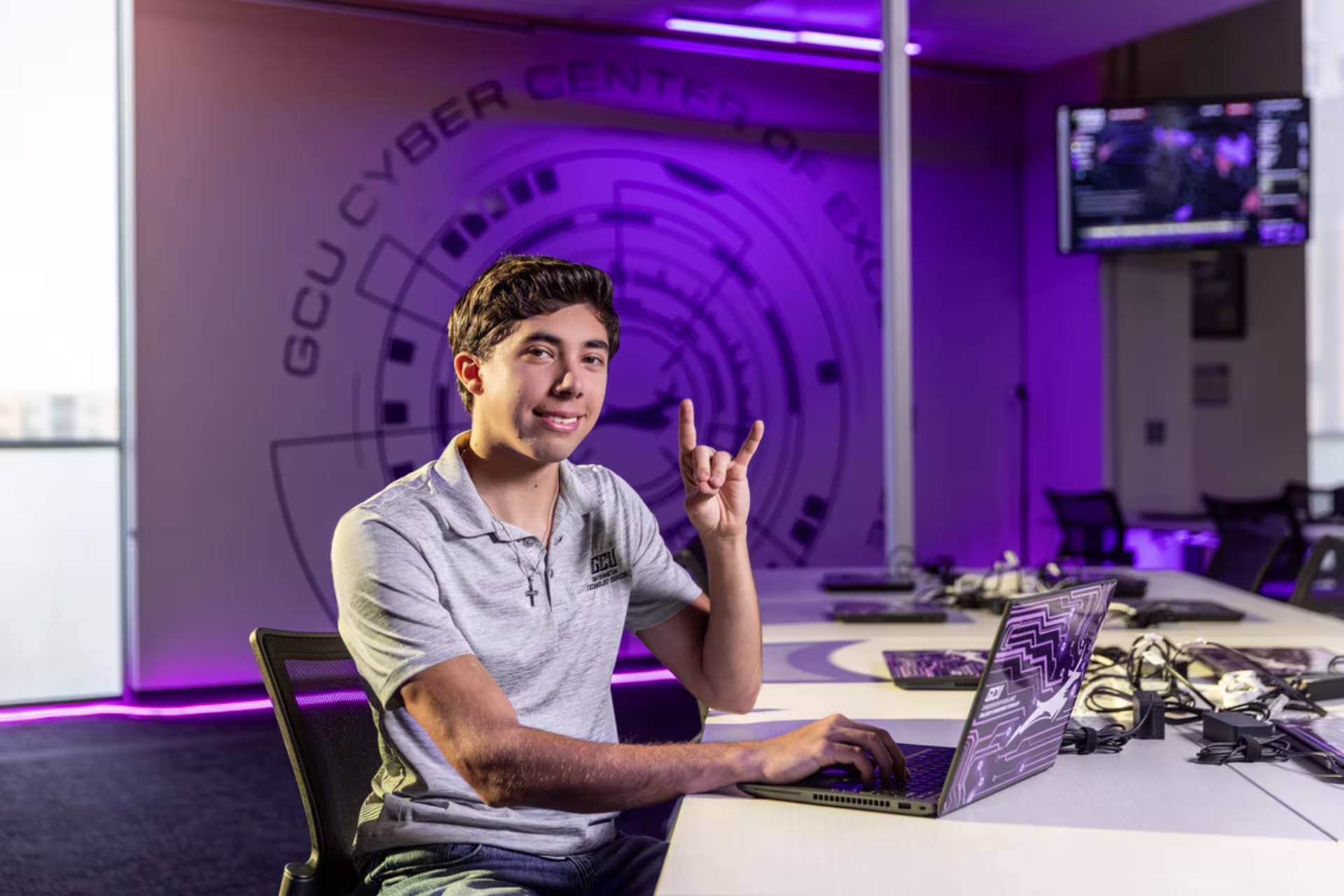 Student sitting at a laptop in a cybersecurity lab with purple lighting and a large circular tech-themed wall graphic, making the lopes up sign and smiling at the camera.