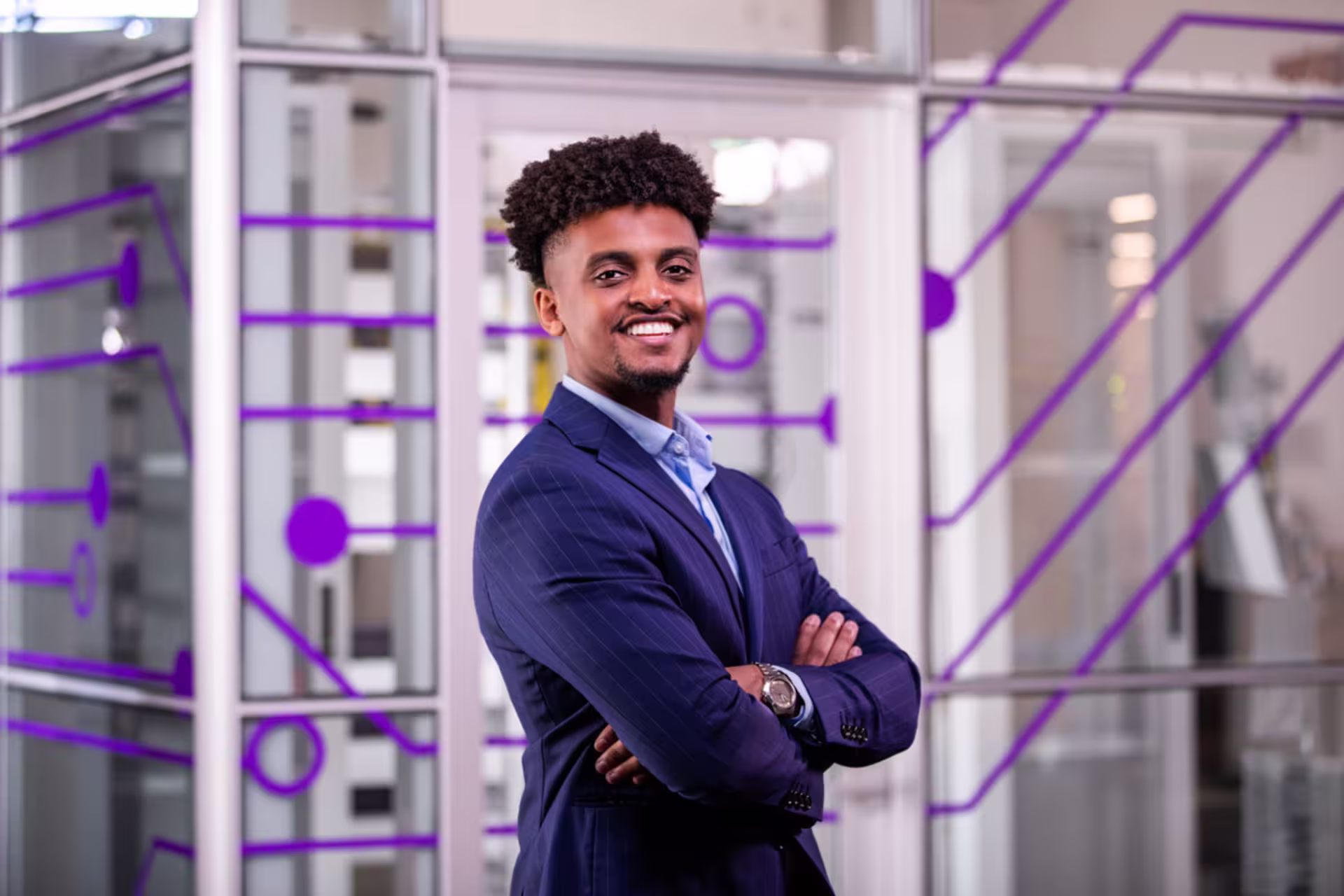 Smiling professional with arms crossed wearing a suit, standing in a modern office space.