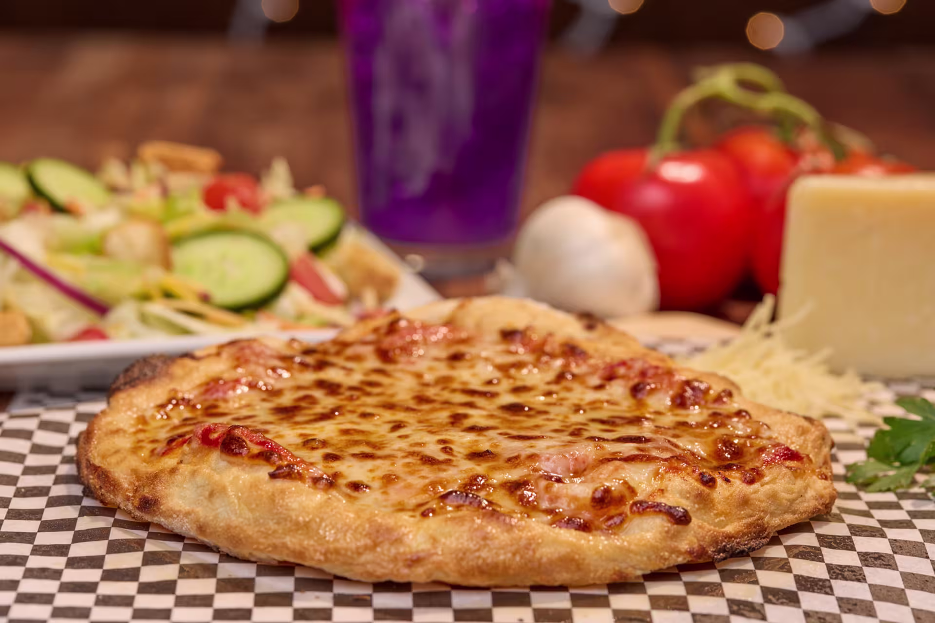Cheese pizza from Havoc House on GCU campus sits on a checkered paper lined tray, with fresh vegetables blurred in the background.