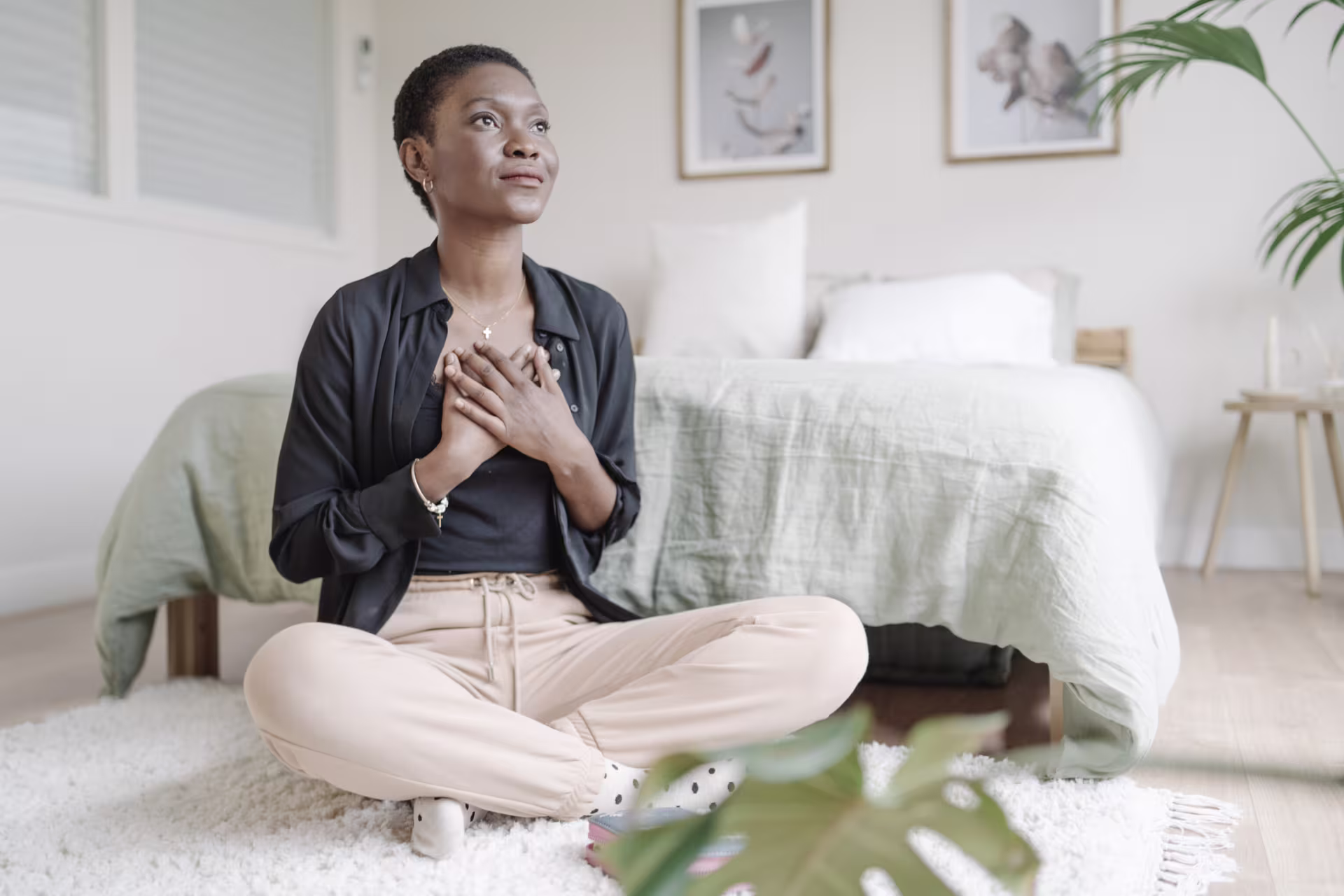 A person sits cross‑legged on the floor of a calm, cozy bedroom with their hands placed over their chest in a peaceful, reflective pose.