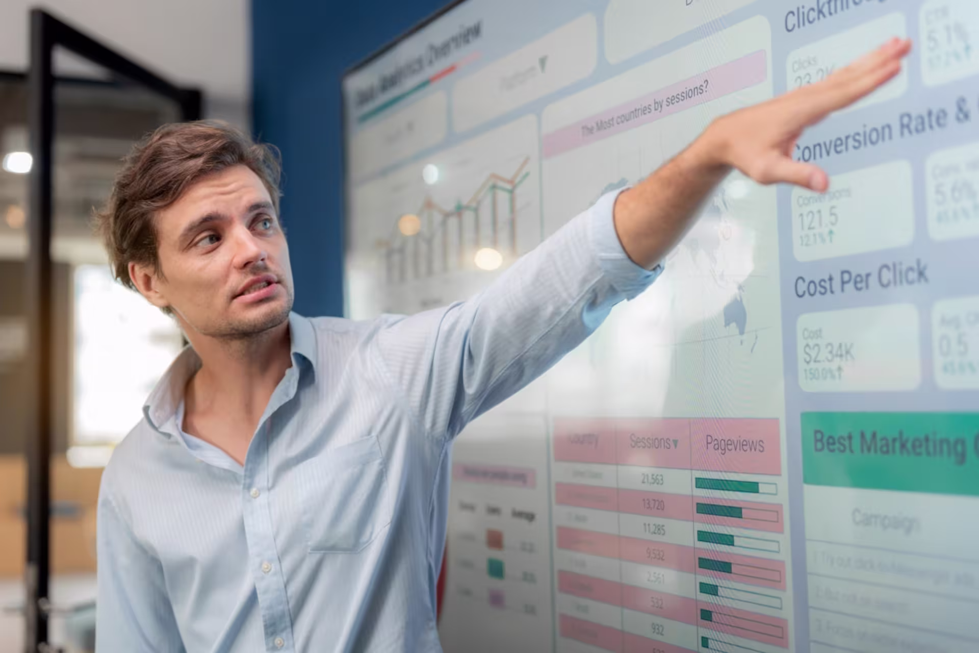 A male operations research analyst points to data visuals on a large screen in an office.