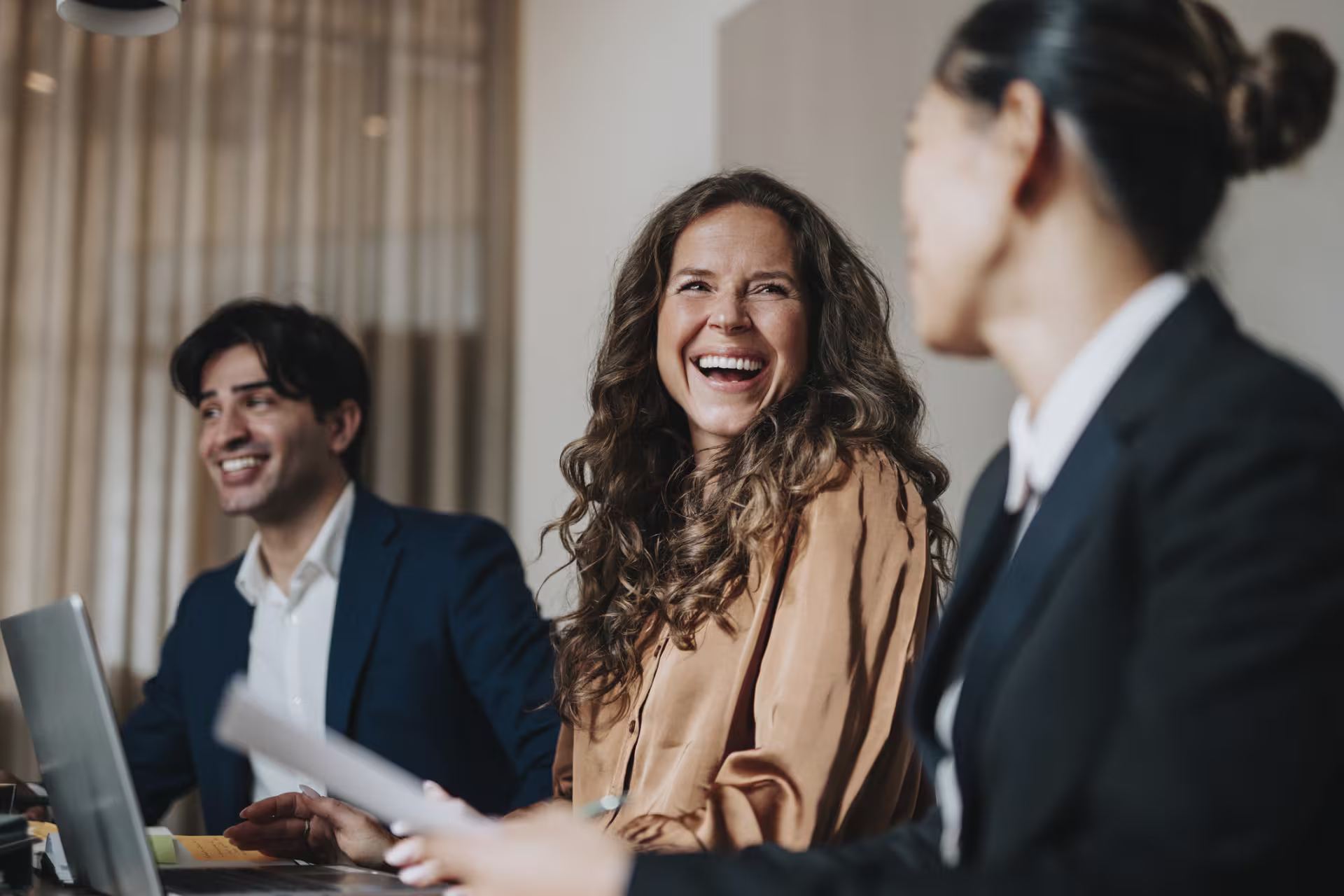 Three business professionals in suits laugh during a meeting at a conference table with a laptop and documents.