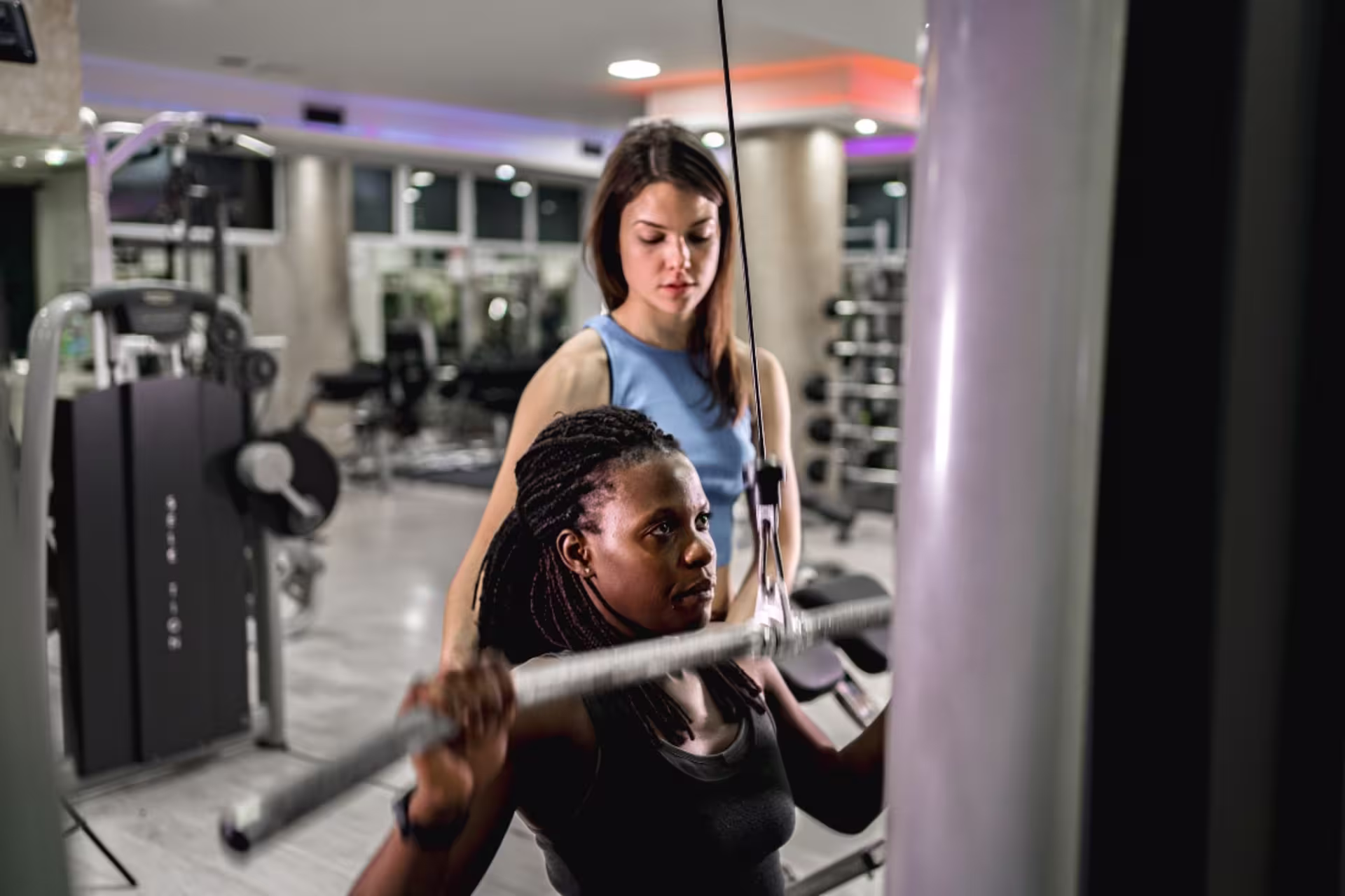 Athletic trainer assisting woman with strength training exercise on lat pulldown machine in fitness center.