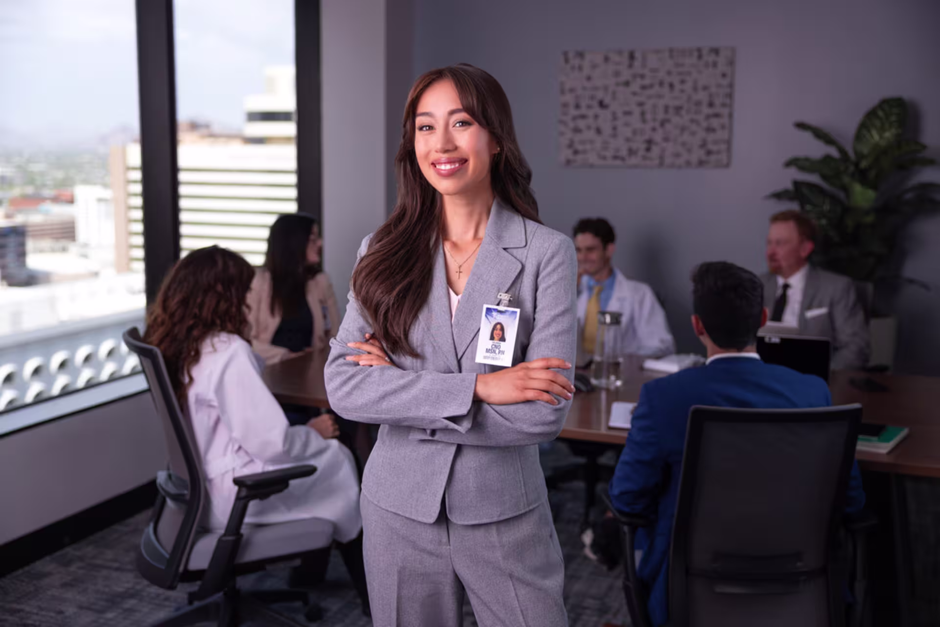 A female FNP professional smiles with her arms crossed while standing in a conference room with other medical staff in the background.