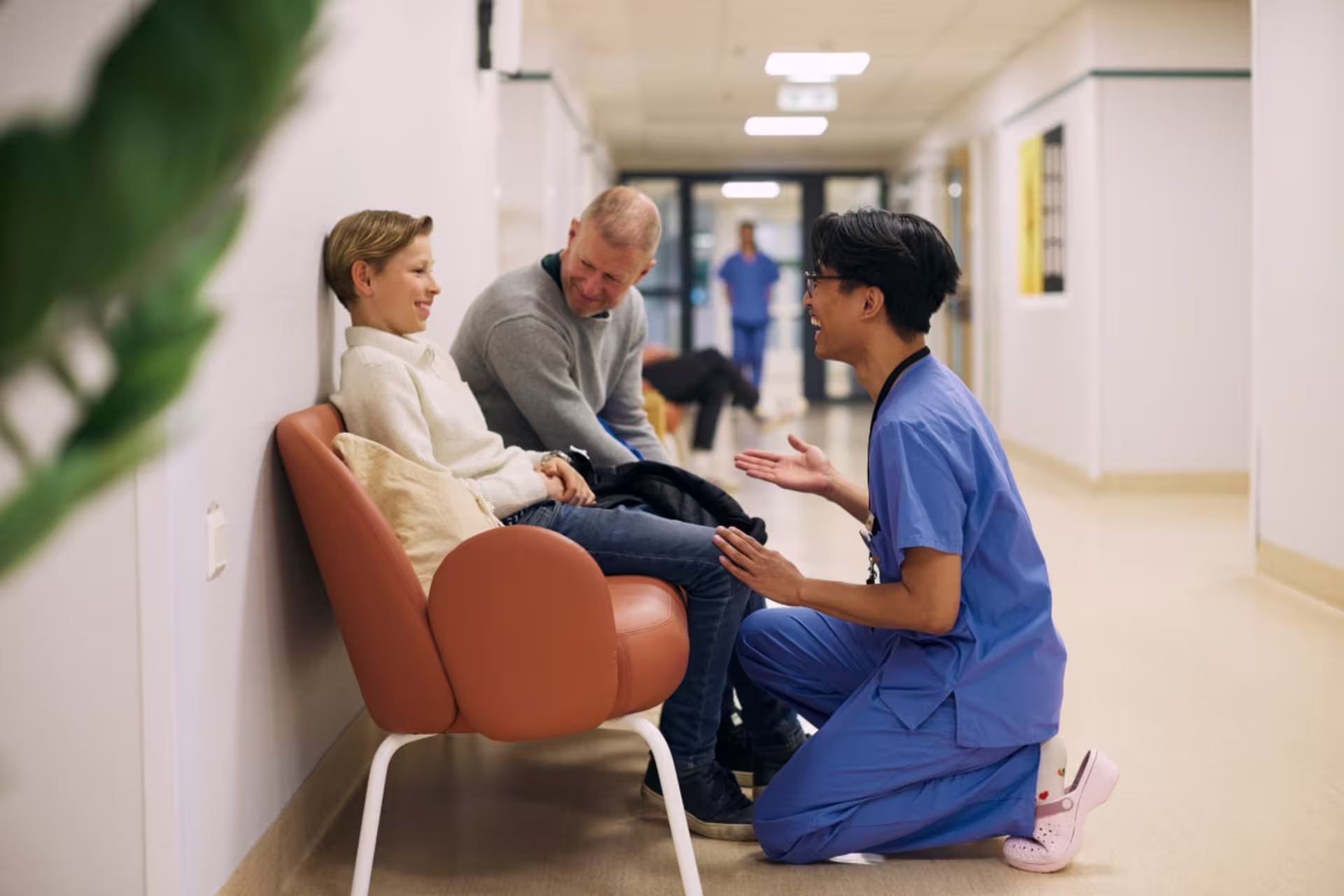 Male nurse smiling while kneeling to speak to a young boy who is seated in a hospital hallway. 