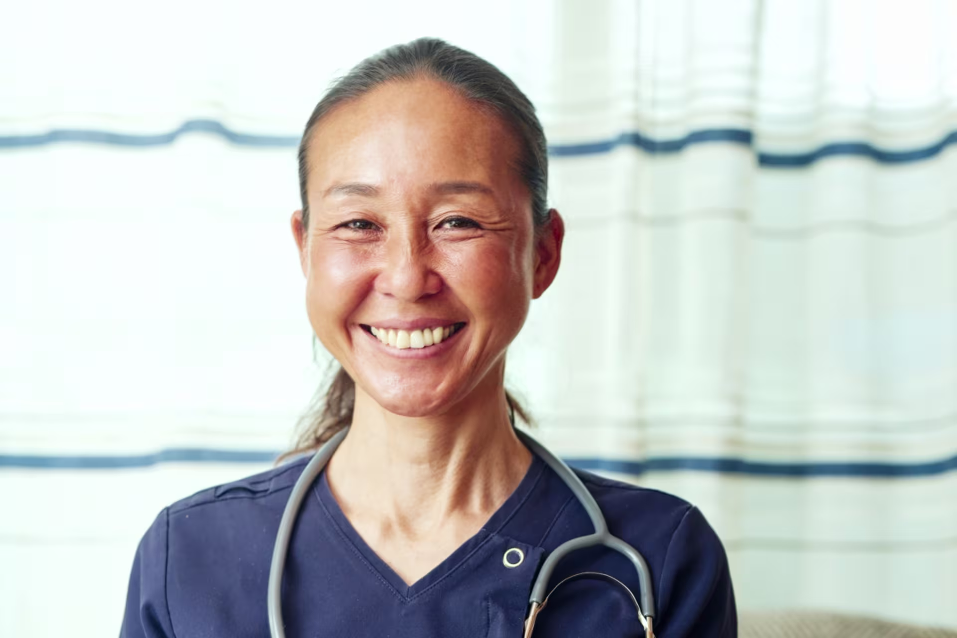 Female nurse stands in front of a hospital curtain, smiling with a stethoscope around her neck.