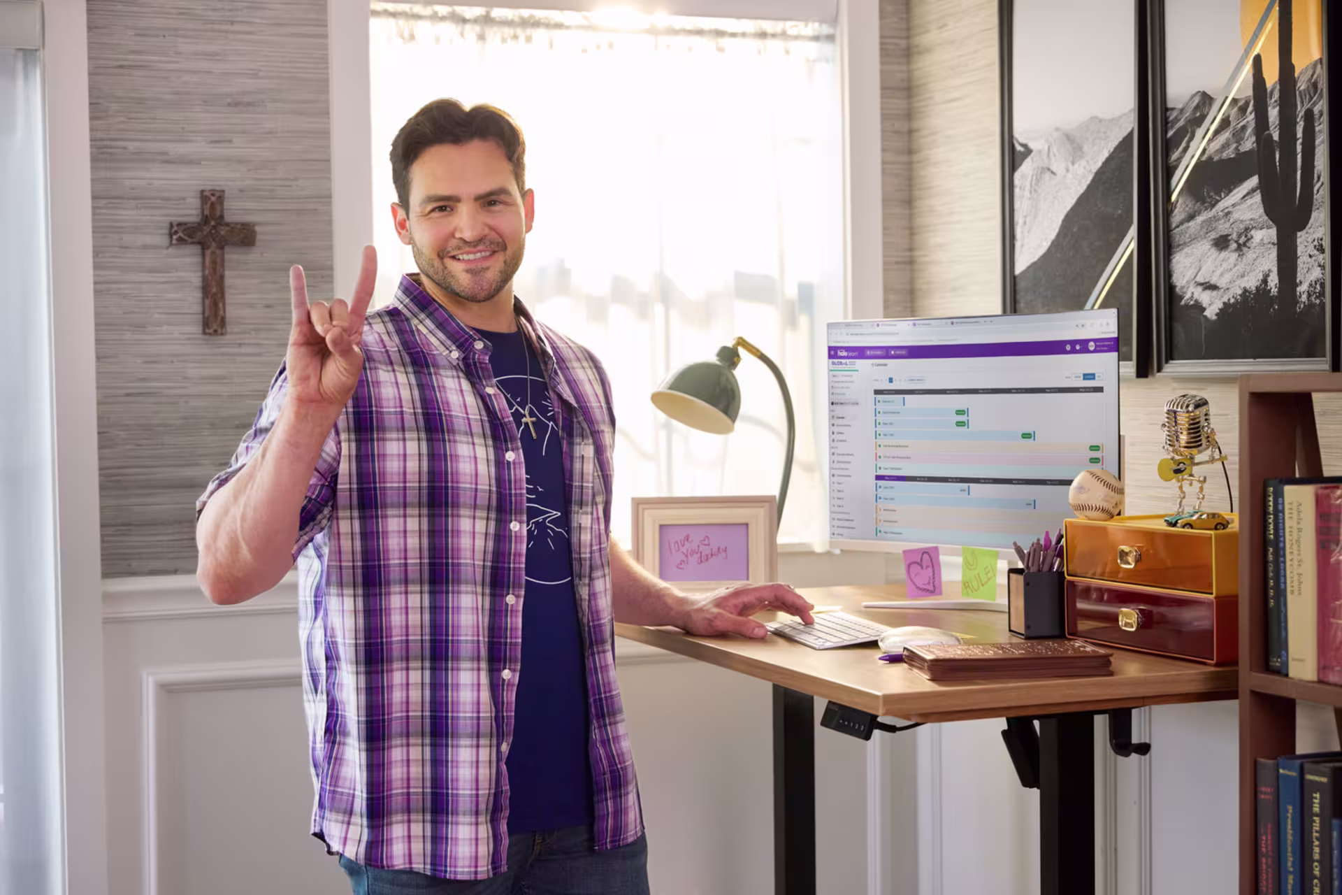 GCU student in plaid shirt giving a Lopes Up hand sign at a standing desk with a monitor displaying an online learning platform.