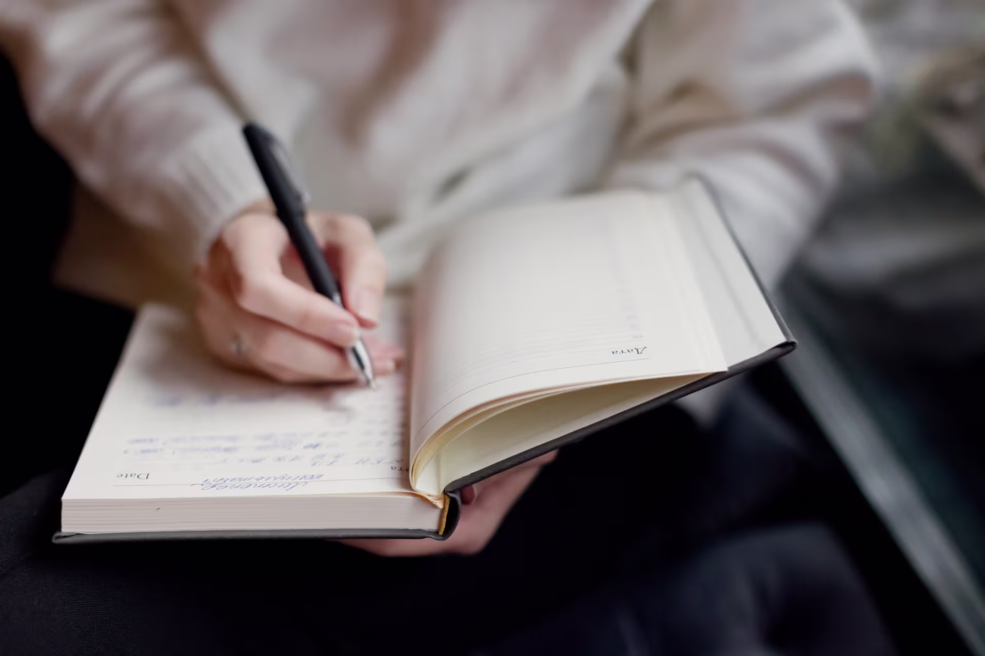 A person writes in an open notebook with a pen, holding the book on their lap.