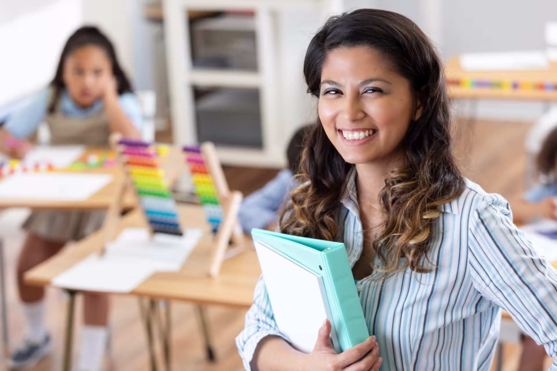 A female elementary education teacher holds a binder while smiling in a classroom, with students seated in the background.