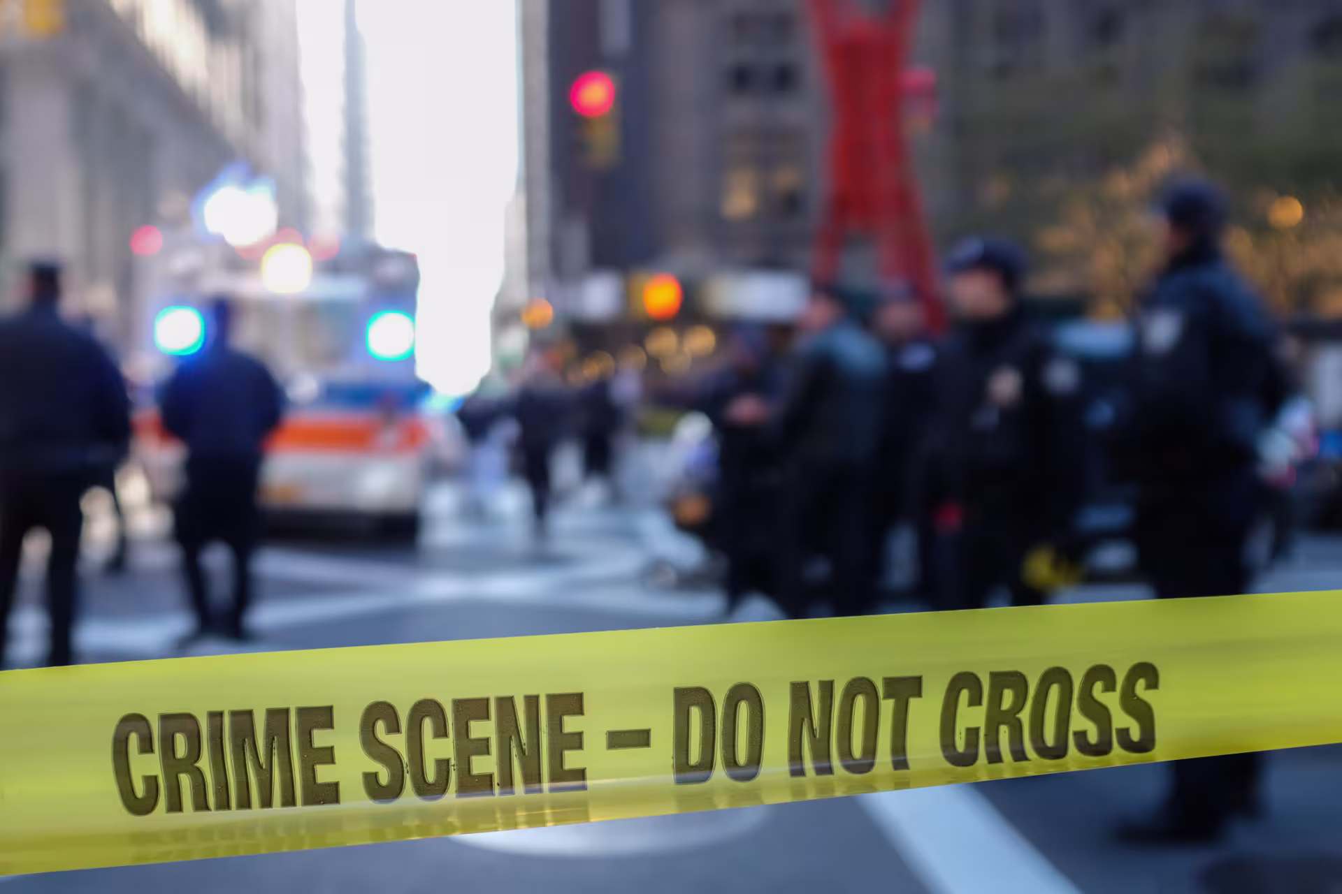 Yellow crime scene tape marked “CRIME SCENE – DO NOT CROSS” stretches across a city street with emergency vehicles and police officers visible in the background.