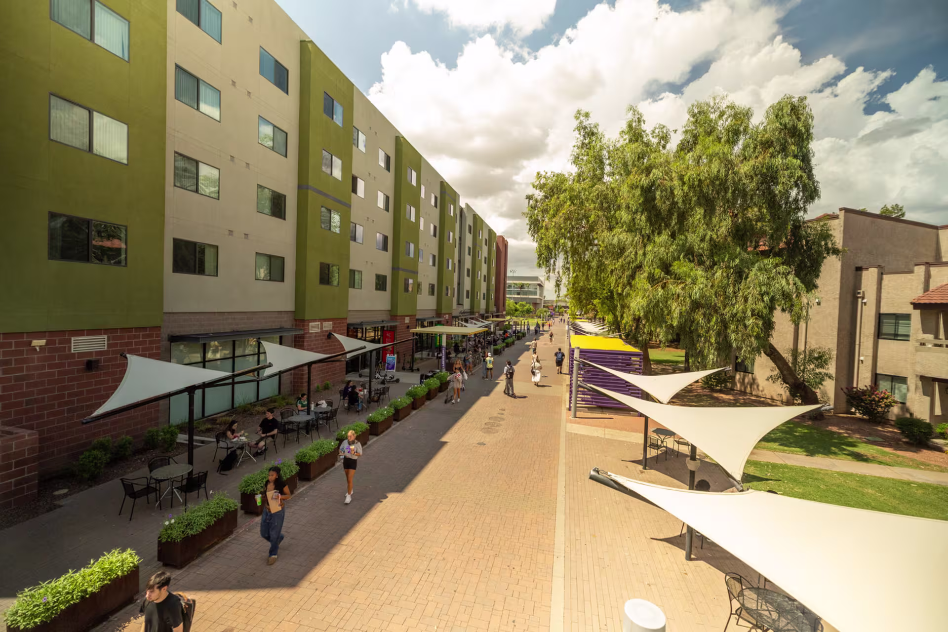Students walking and dining along Lopes Way at GCU with outdoor seating, shade structures and campus buildings on a sunny day.