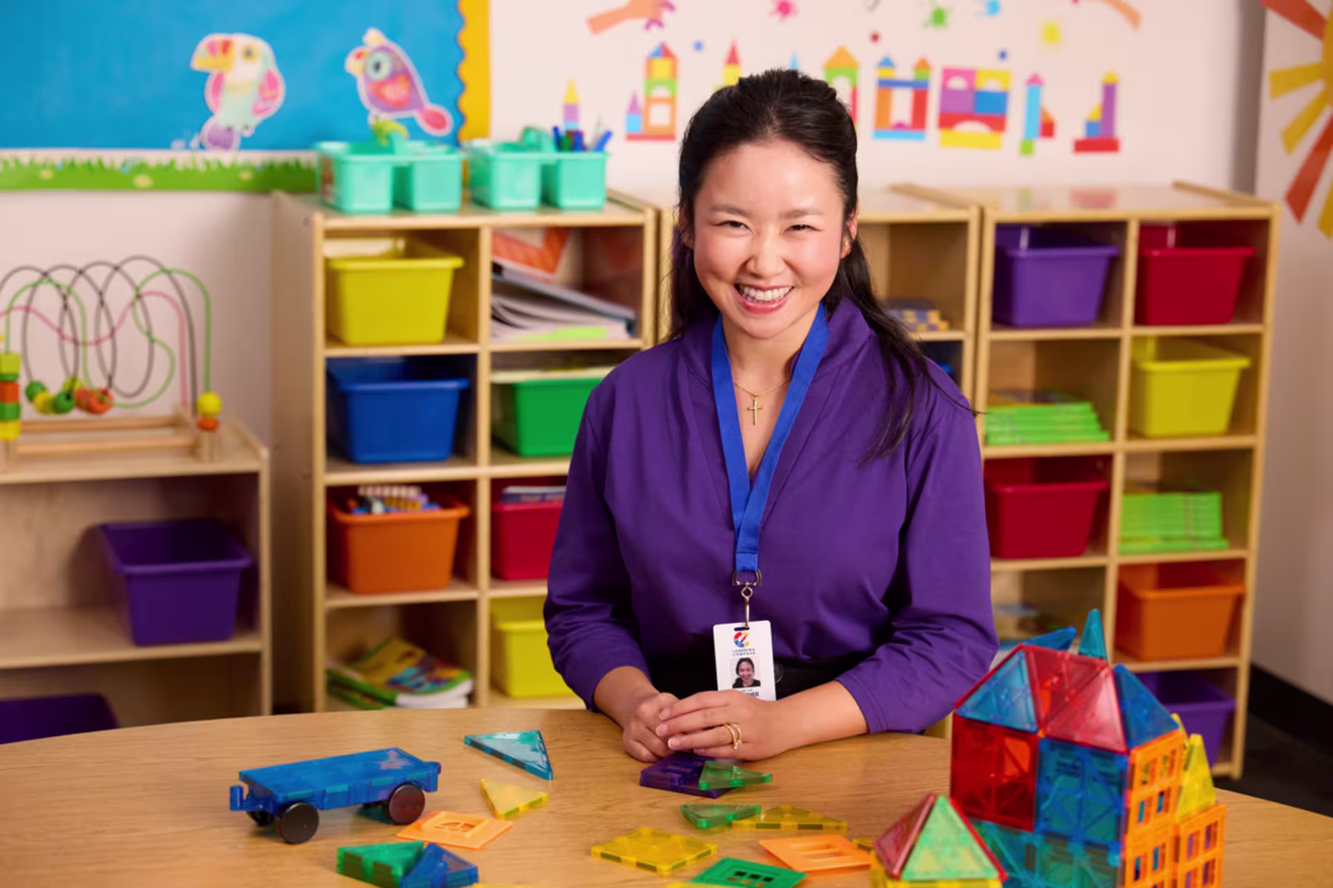 A female undergraduate teaching student sits at a classroom table with colorful educational toys and storage bins in the background.