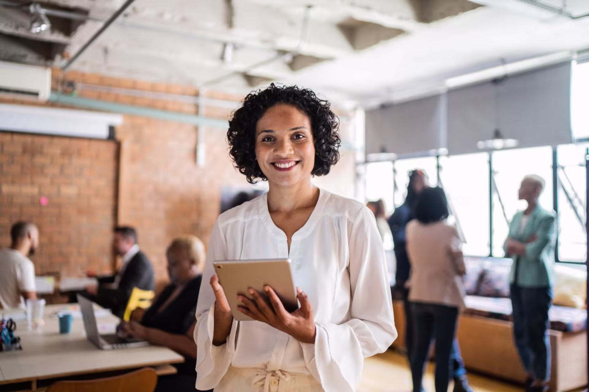 A female business professional holds a tablet while standing in a collaborative office workspace.